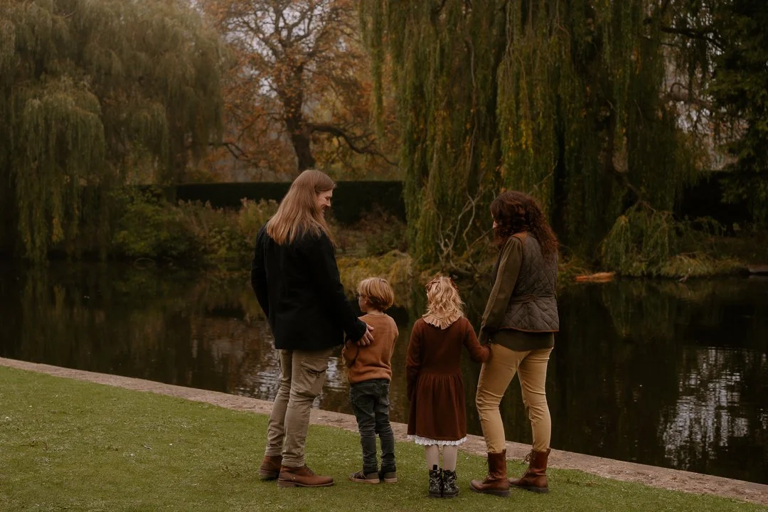 Two women and two children holding hands by a river in a park during fall.