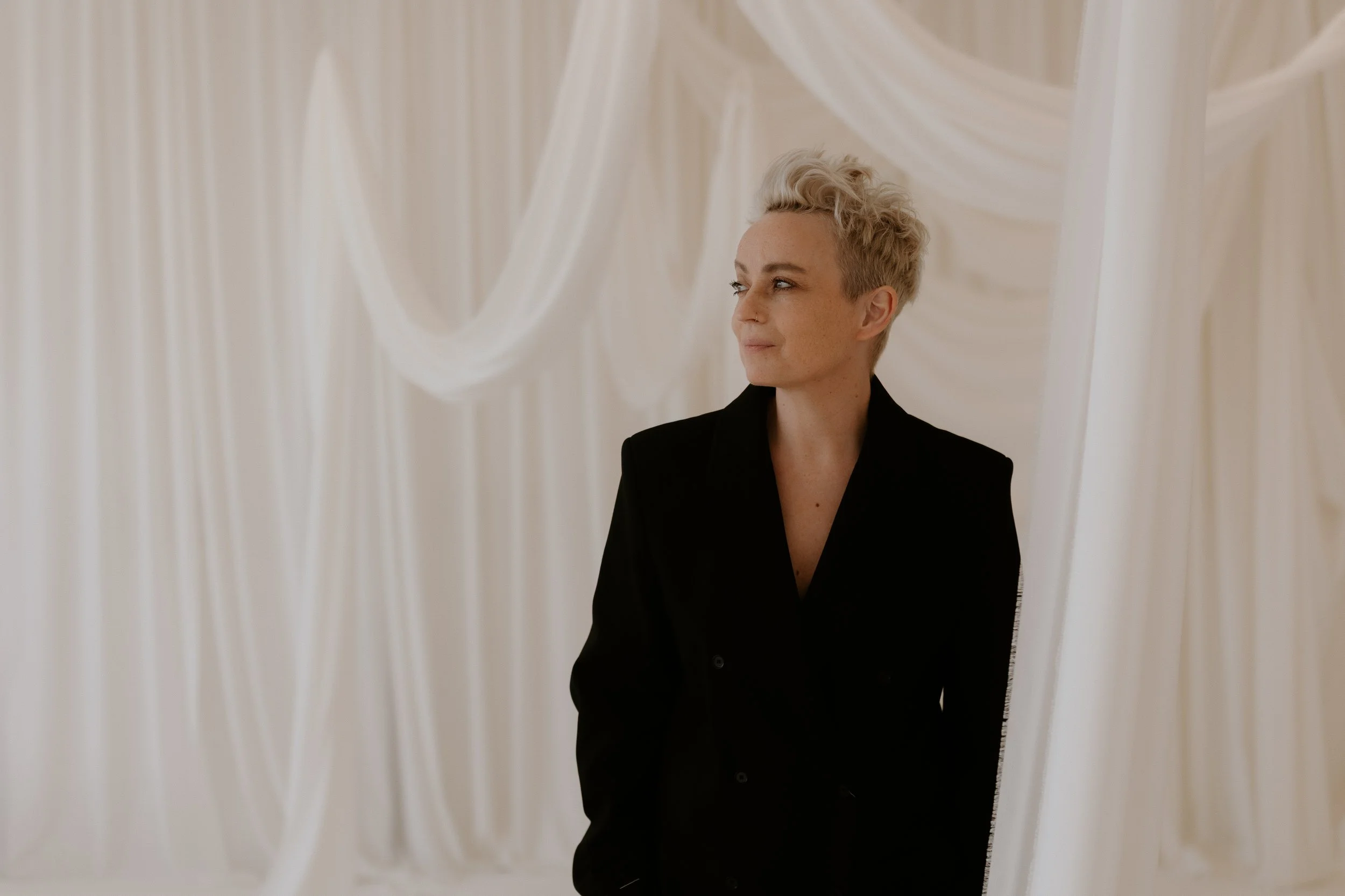 A person with short blond hair and a black shirt standing in front of a white, draped fabric backdrop.