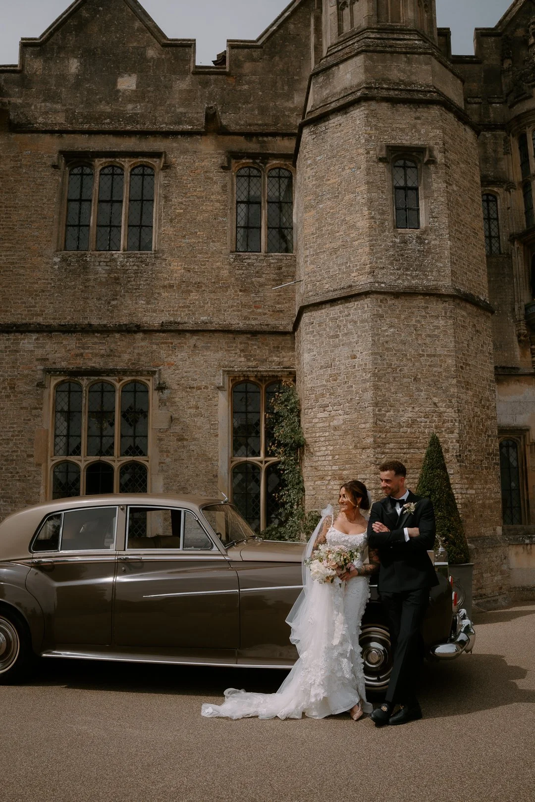 Bride and groom in wedding attire standing beside a vintage car in front of an old stone building with tall, arched windows.