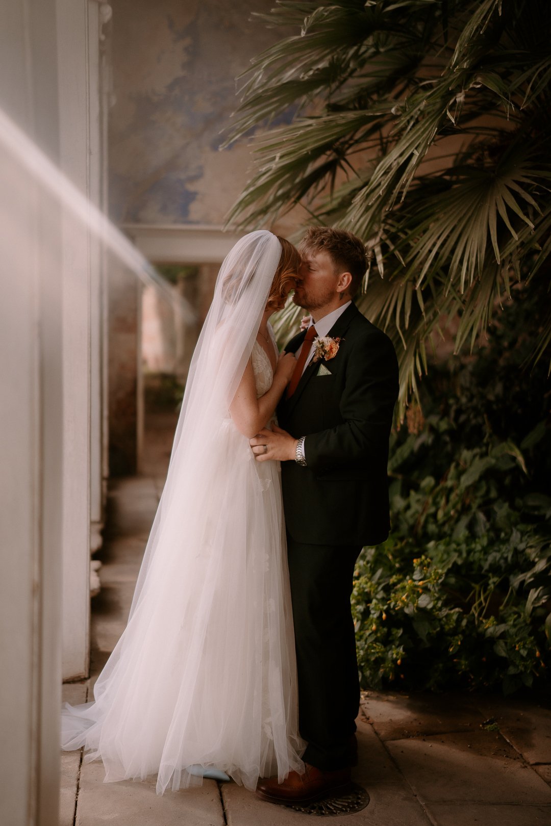 A bride and groom sharing a kiss at their wedding, standing outdoors near greenery and tropical plants.