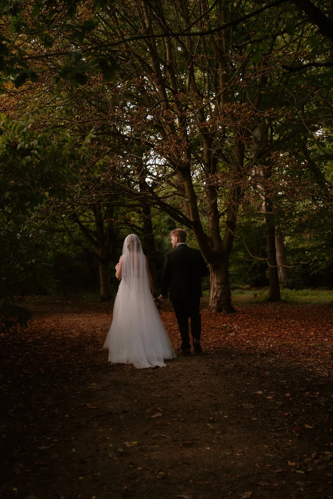 A bride and groom walking hand in hand through a wooded area, with tall trees and fallen leaves on the ground, during dusk or early evening.