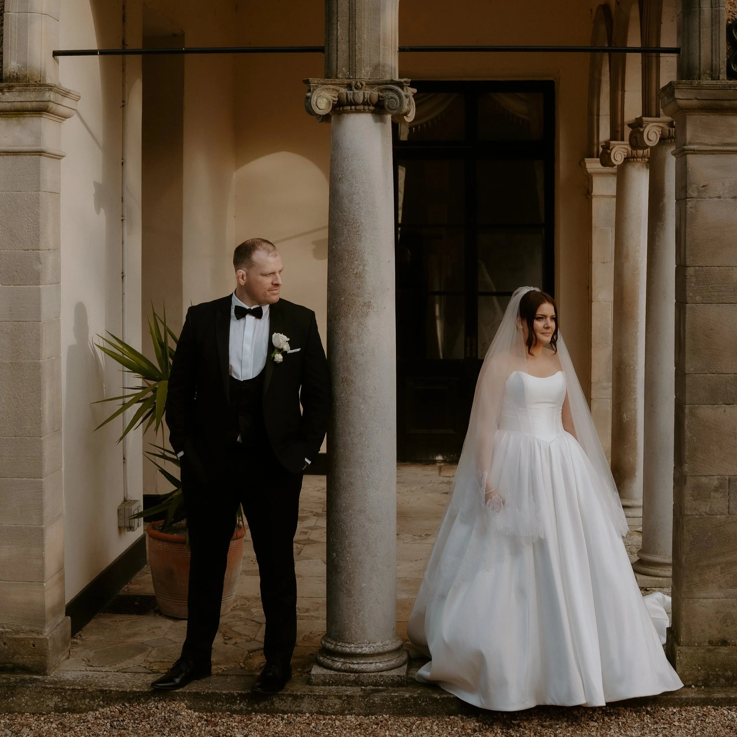 Half of a man in a tuxedo and half of a woman in a wedding dress standing outdoors under an arched stone structure.