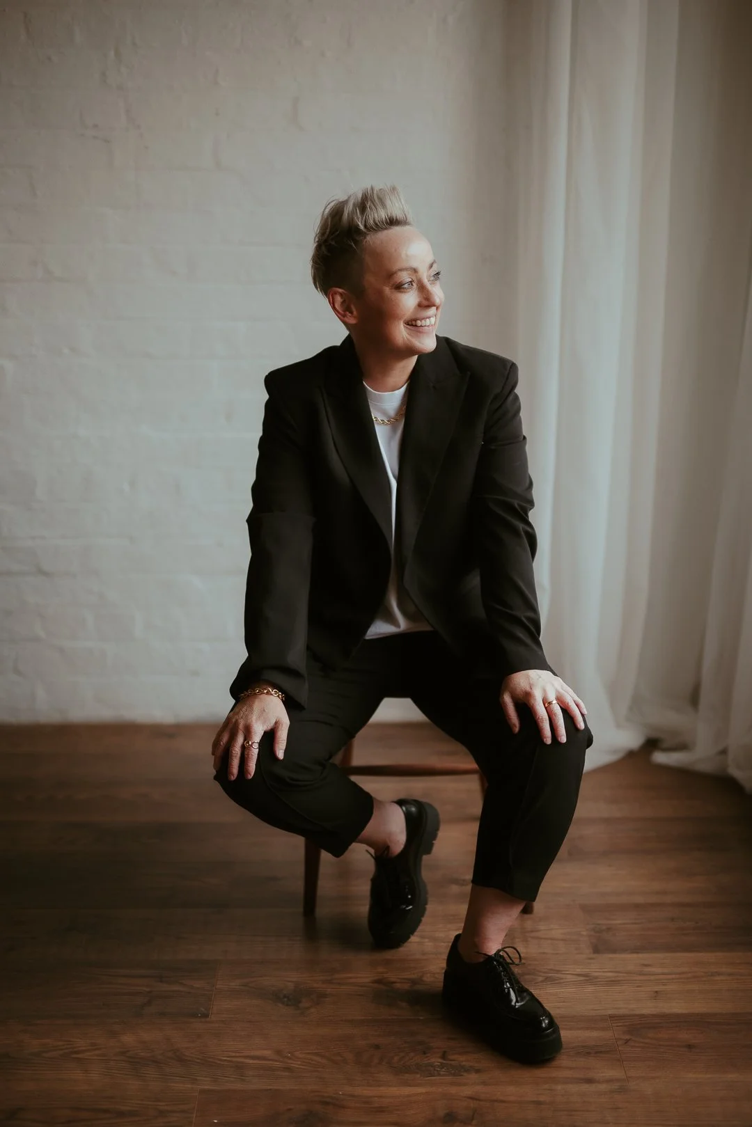 Woman with short blonde hair wearing a black suit sitting on a chair, smiling and looking to the side, in a room with wooden floors and white brick wall.