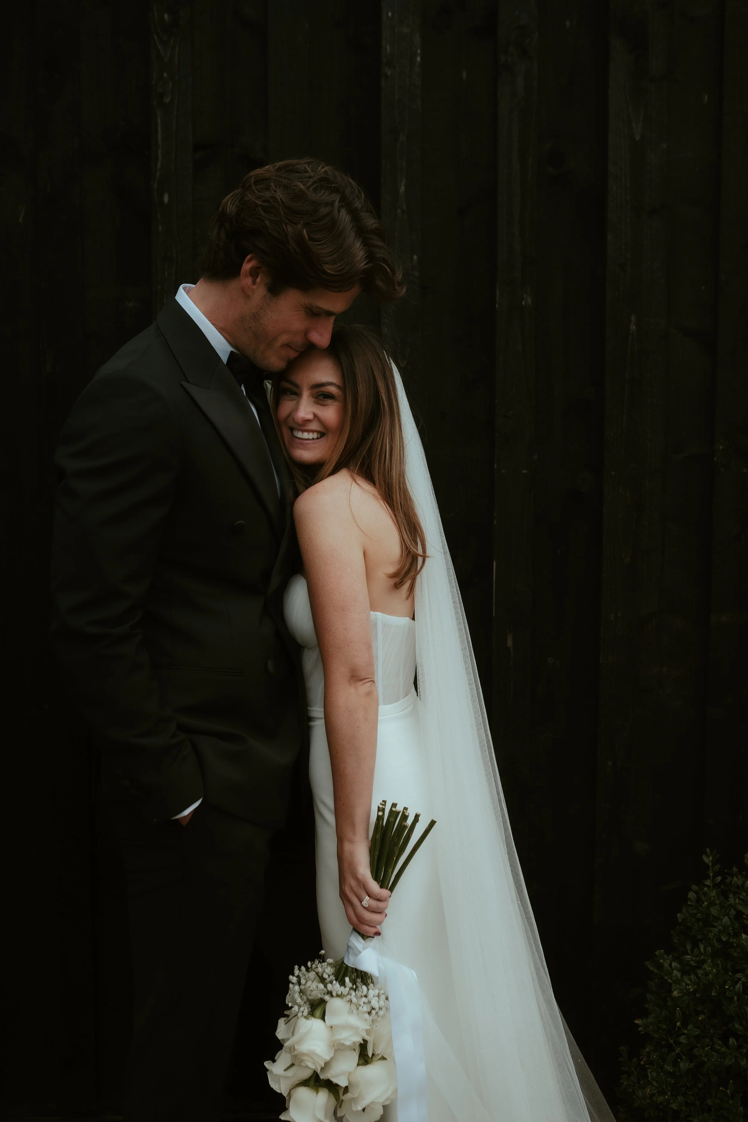 A bride and groom standing close together, smiling, against a dark wooden background. The bride is holding a bouquet of white flowers and wearing a strapless white wedding gown with a veil. The groom is dressed in a black tuxedo with a bowtie.