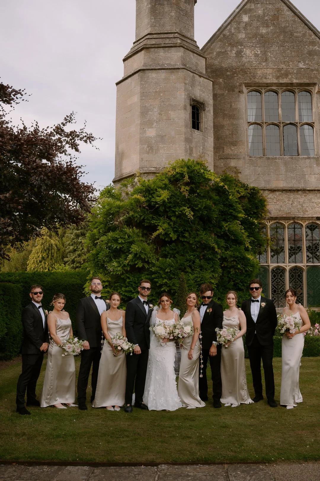 A group of wedding party members, dressed in formal attire, standing outside of a historic stone church surrounded by greenery, during daytime.