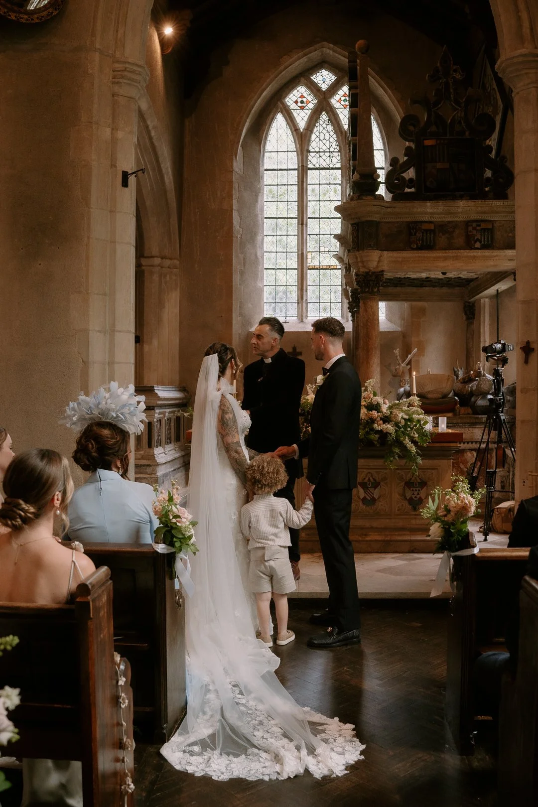A wedding ceremony taking place inside a church, with a bride and groom holding hands before an officiant, surrounded by seated guests and floral decorations.
