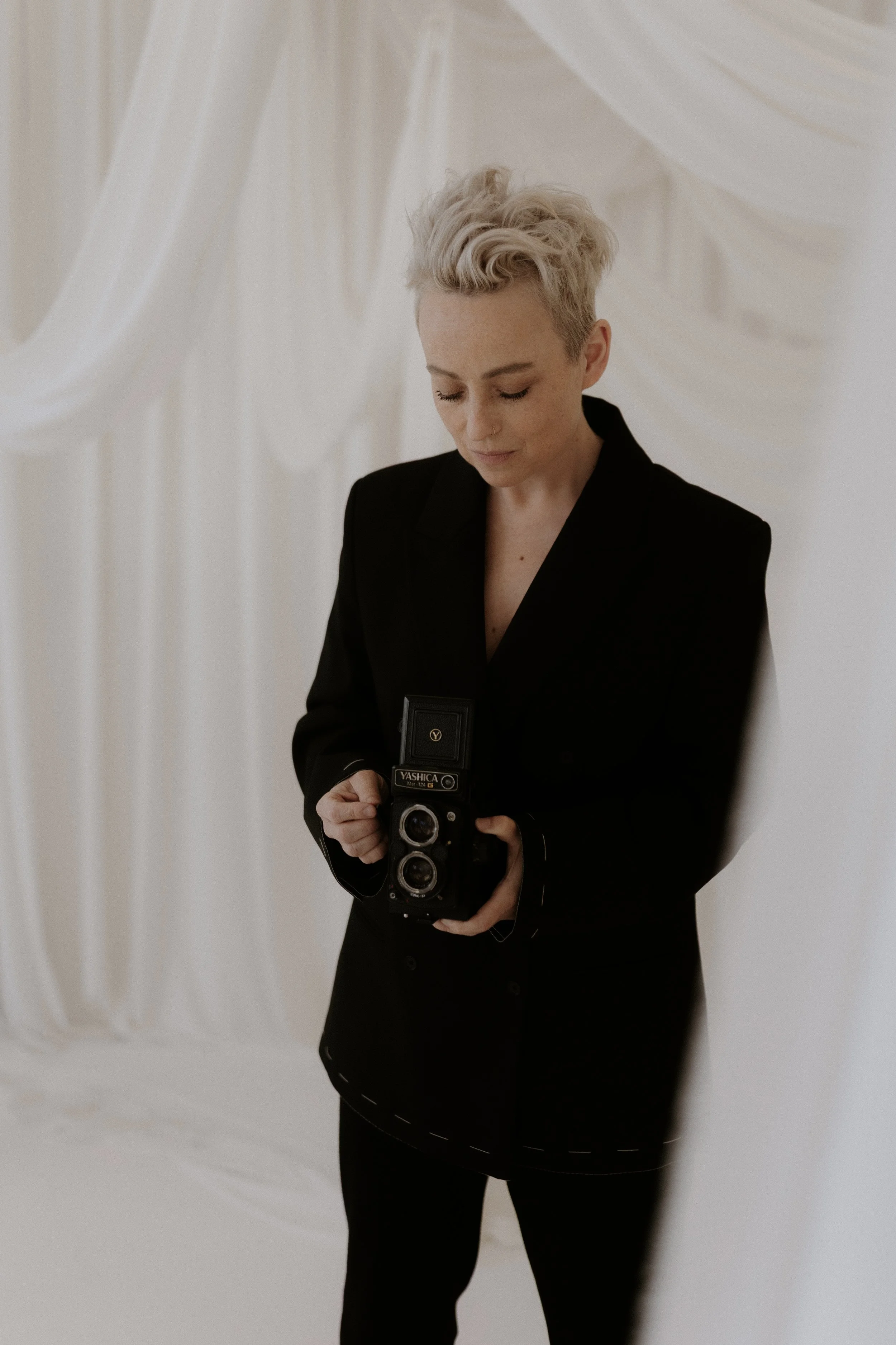 A person with short, platinum blonde hair holds a vintage camera while standing in front of draped white fabric.