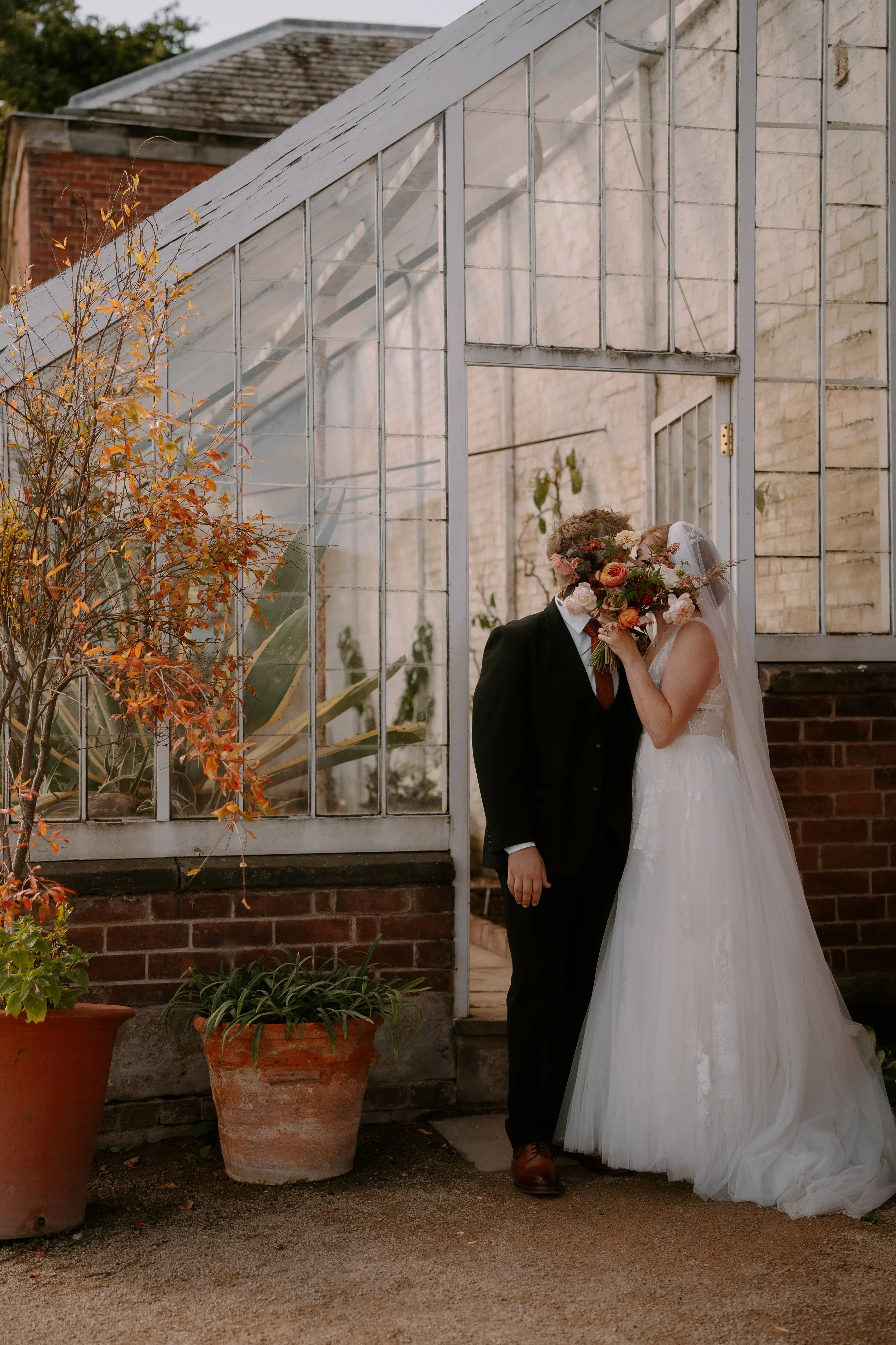 Bride and groom sharing a kiss outdoors, with the bride holding a large bouquet, standing in front of a glass greenhouse.