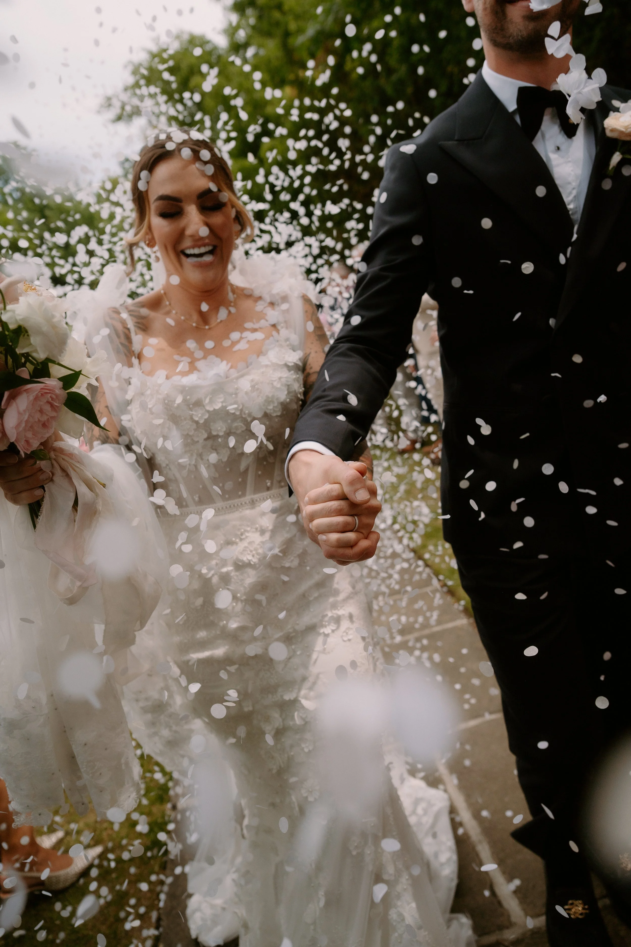 A bride and groom holding hands at a wedding, surrounded by falling white confetti, with the bride smiling and wearing a white wedding dress and holding a bouquet of flowers.