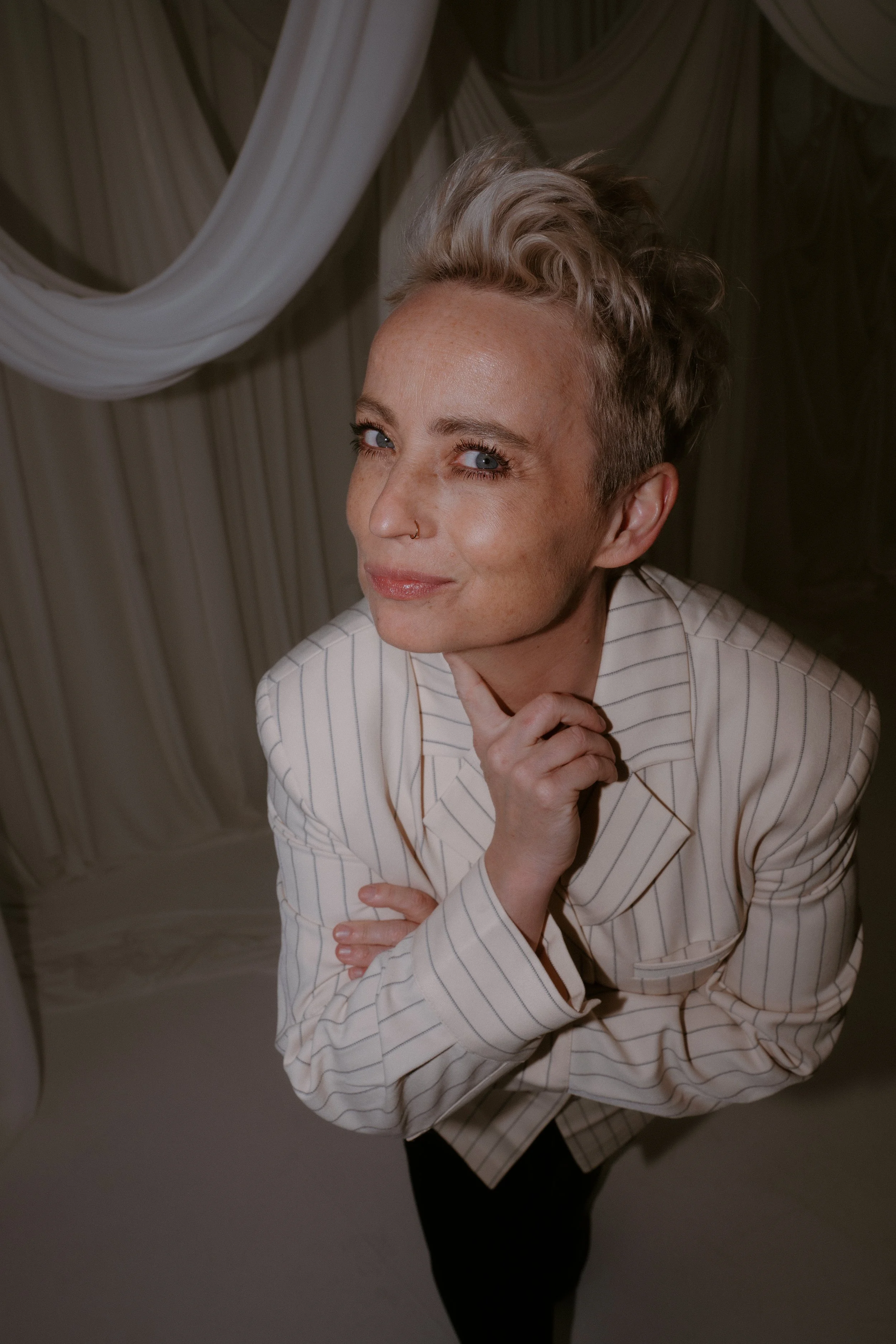 A woman with short, styled blonde hair and a nose piercing, wearing a white pinstripe blazer, is posing with her left hand resting under her chin. She is smiling slightly and looking upward. The background features draped fabric in neutral tones.