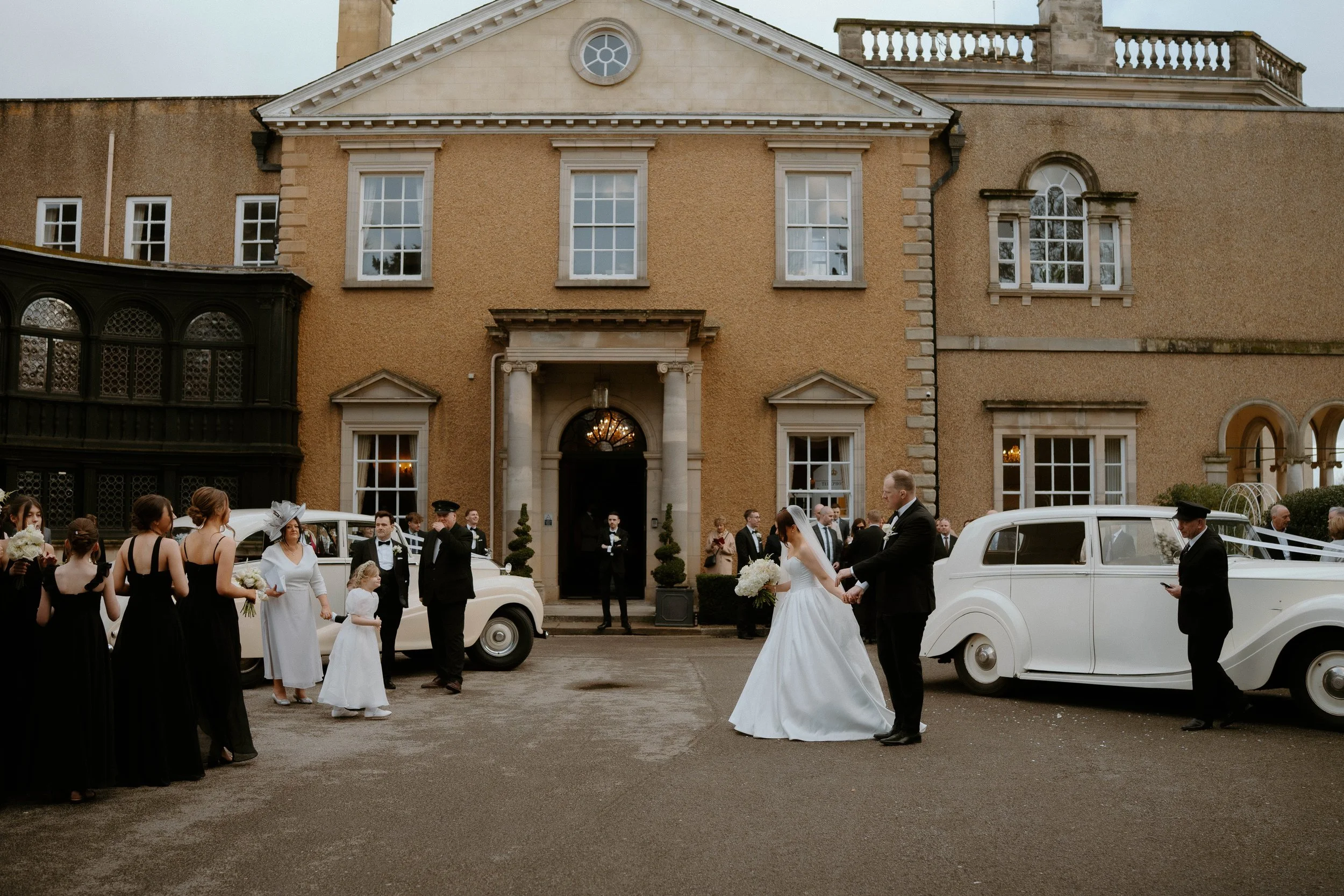 Bride and groom holding hands outside a large historic house, surrounded by wedding guests, vintage cars, and attendants in formal attire.