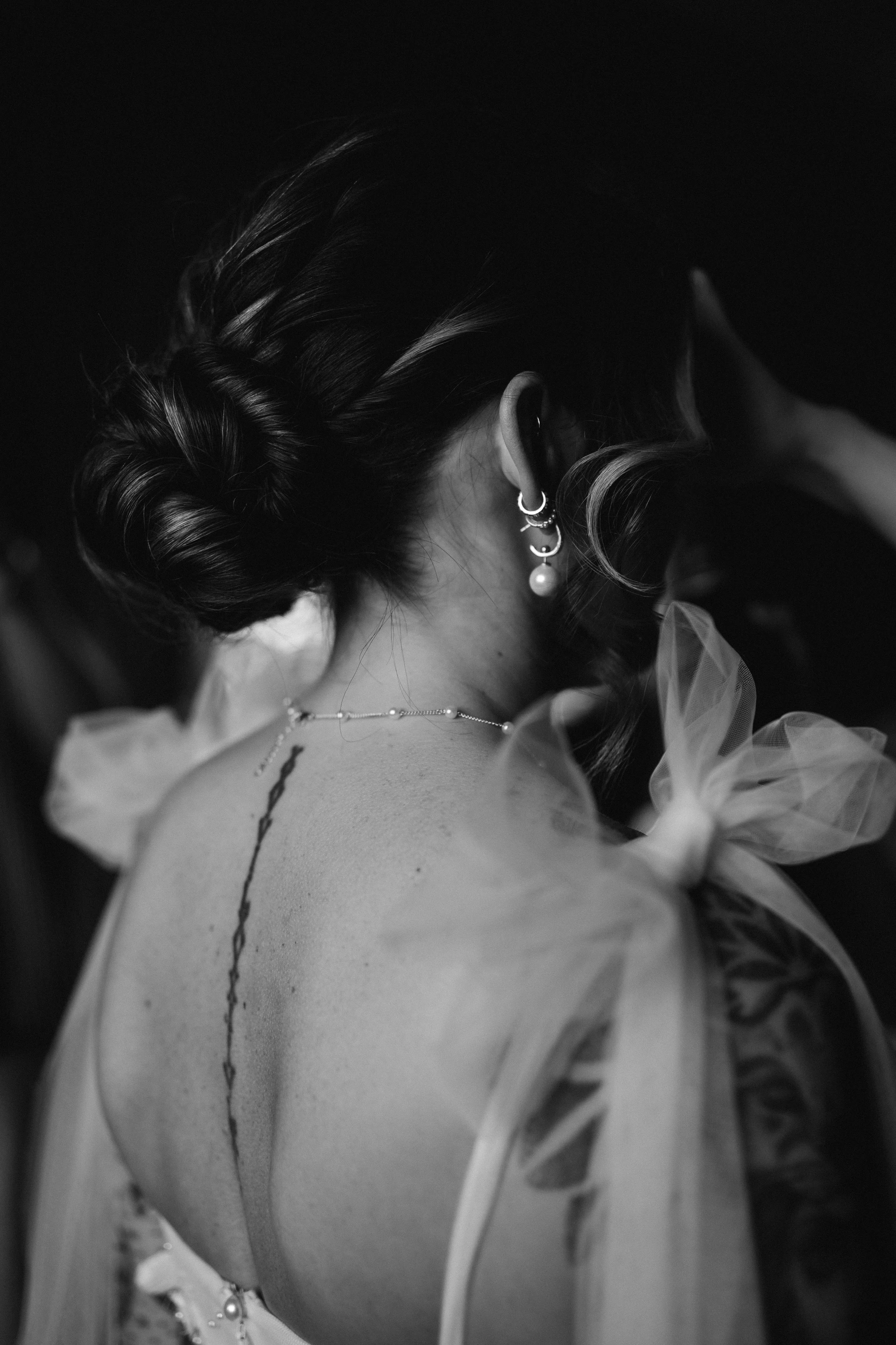 Close-up of a person's partially visible face, ear with multiple earrings, braid hairstyle, necklace, and a tattoo on shoulder, in black and white.