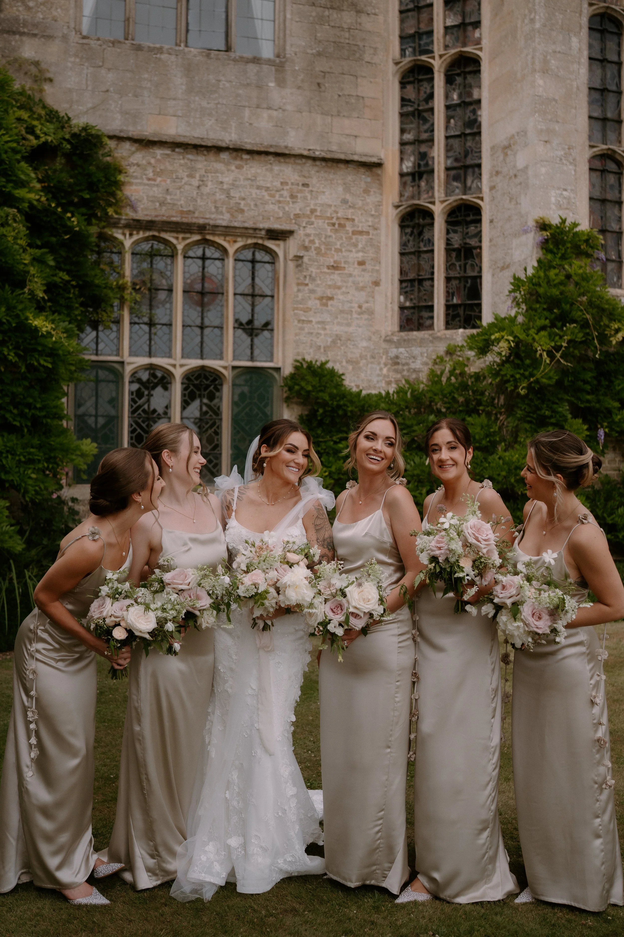 A group of women dressed in matching beige satin bridesmaid dresses, holding bouquets of pink and white flowers, standing outside in front of an old stone building with large stained glass windows, smiling and posing for a picture.