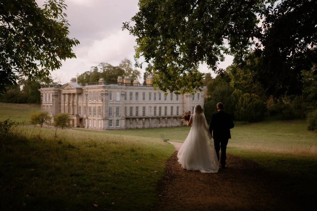Bride and groom walking towards a large historic mansion on a dirt path through a park with trees.