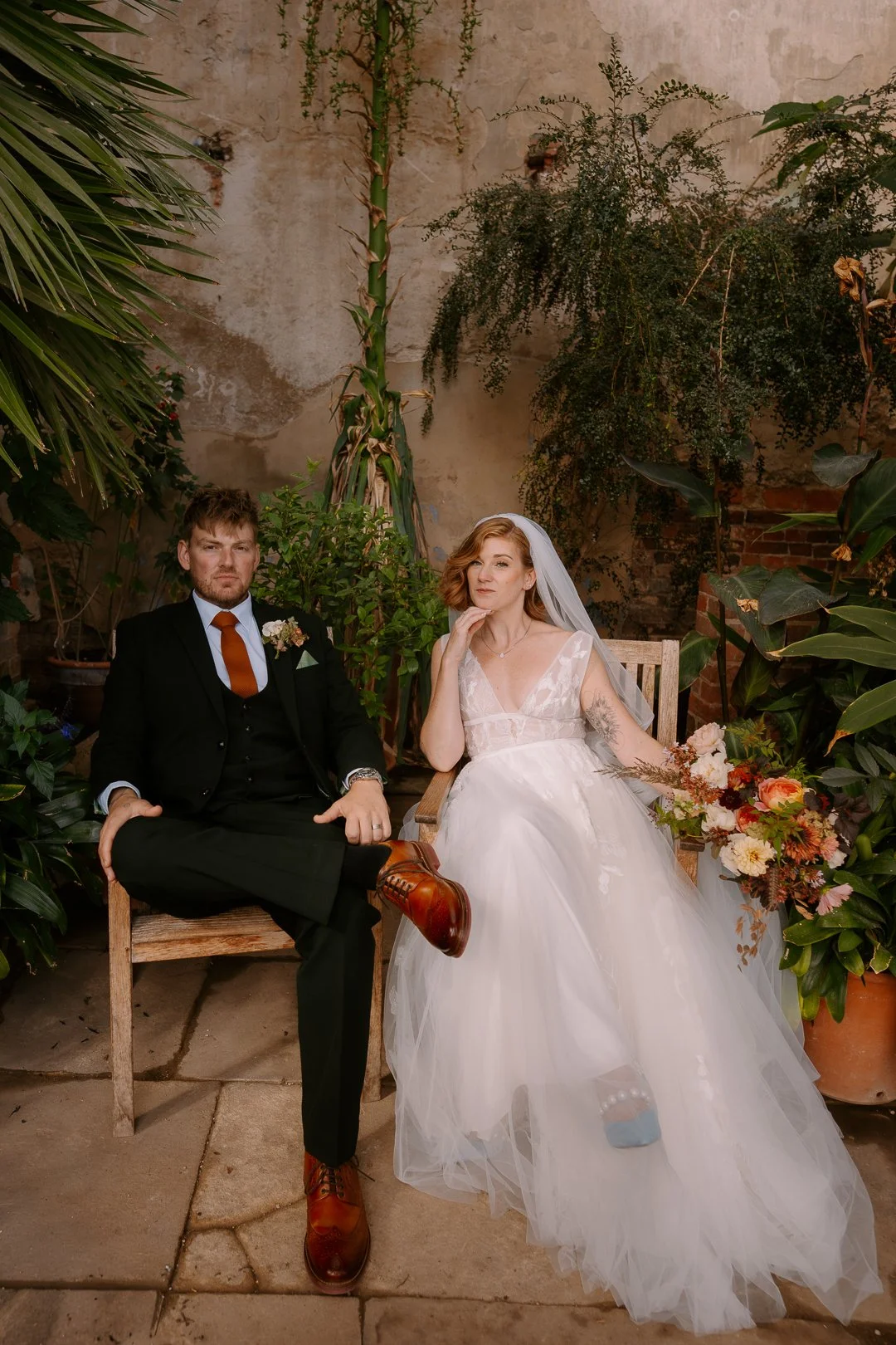 A bride and groom seated outdoors among greenery and plants, with a weathered wall in the background. The groom is wearing a black suit with a white shirt, orange tie, and brown shoes, while the bride is wearing a white wedding dress with lace detail