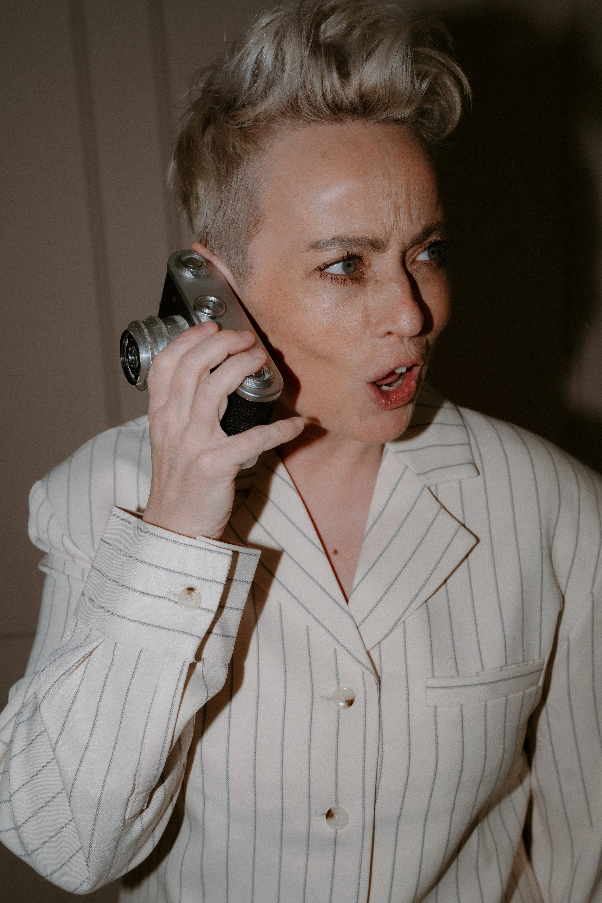 A woman with short, blonde hair styled in a pompadour, wearing a white pinstripe shirt with brown piping, holds a vintage film camera to her ear as if making a phone call, with an intense and expressive facial expression.