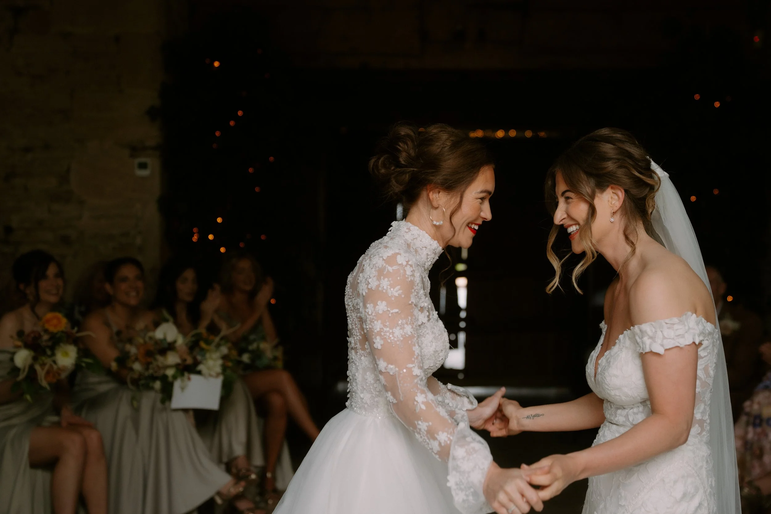 Two women, dressed in wedding gowns, face each other holding hands during their wedding ceremony at night. In the background, several women in matching dresses sit with bouquets, smiling and observing.