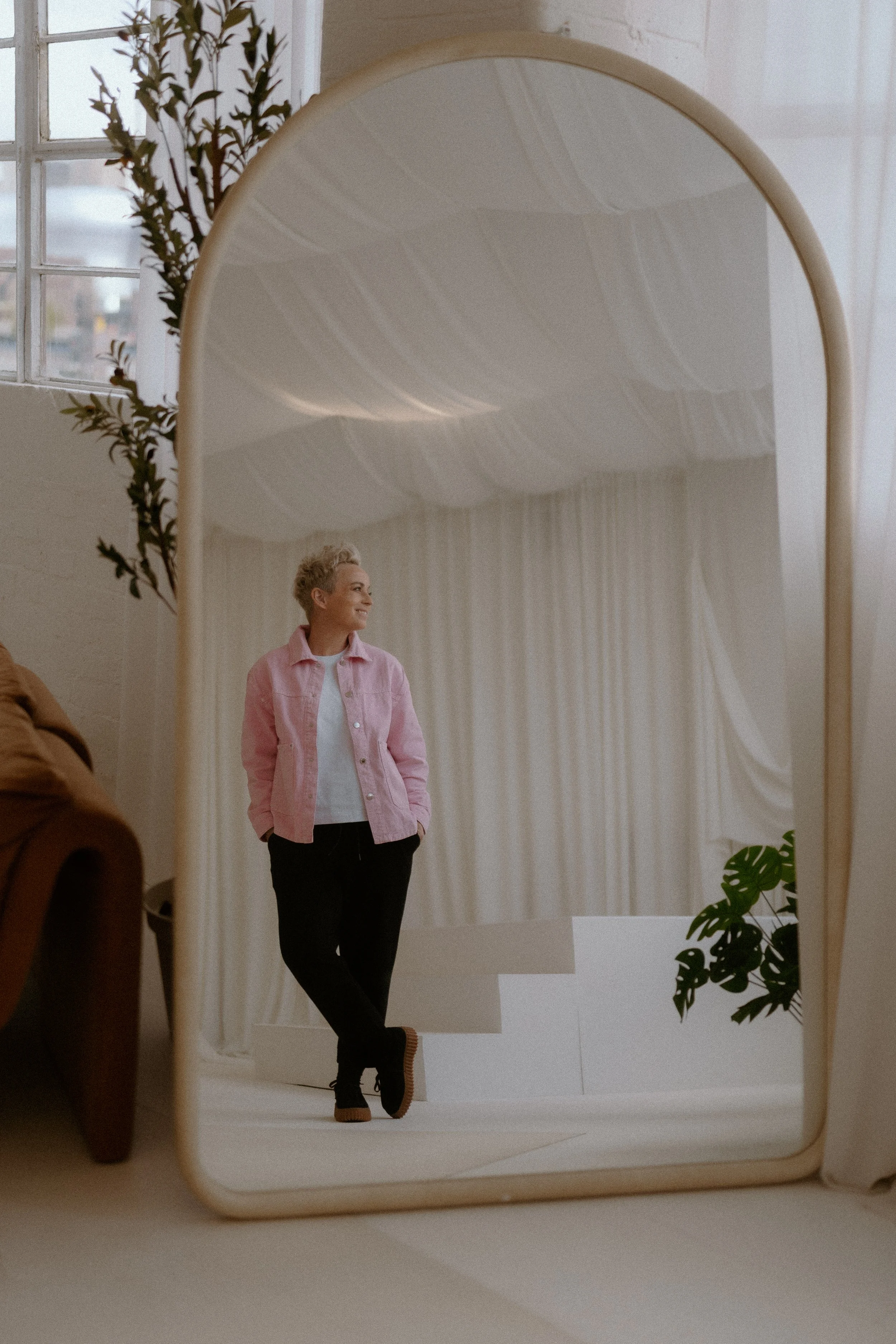 A woman with short blonde hair wearing a pink jacket and black pants standing in front of a large, curved mirror, smiling and looking to her right. The reflection shows a room with striped beige and white curtains and a white wall, with some green plants visible at the bottom right corner.