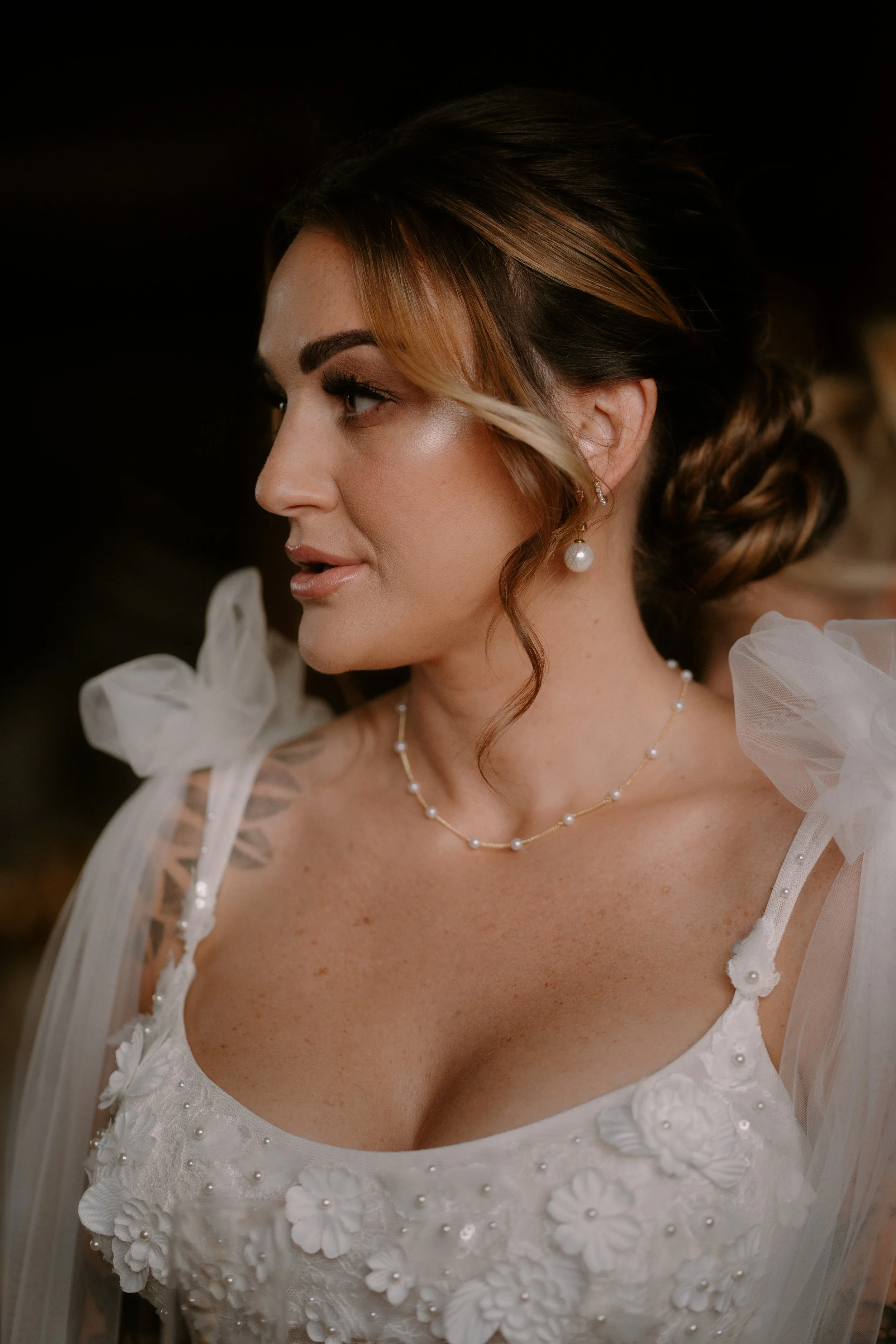 Close-up of a woman with styled brown hair, wearing pearl jewelry, and a white dress with floral lace details.