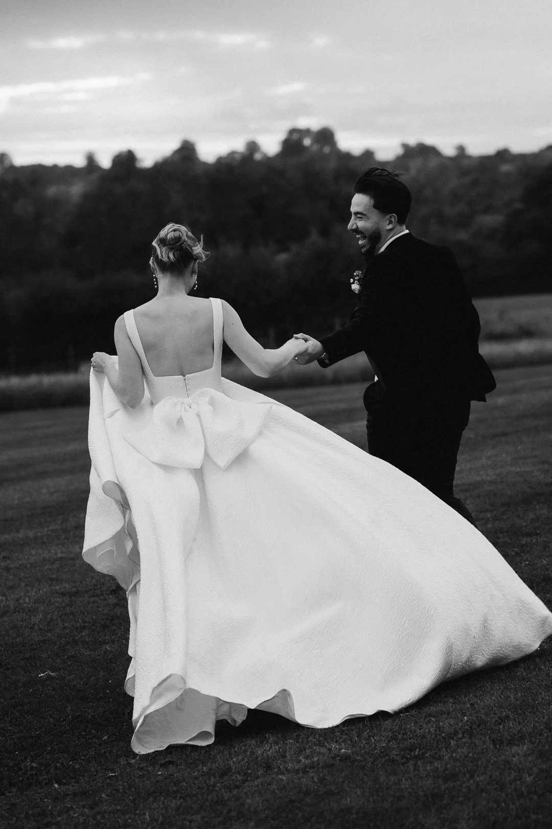 A bride and groom holding hands and dancing outside on a grassy field during their wedding, with trees and a cloudy sky in the background.
