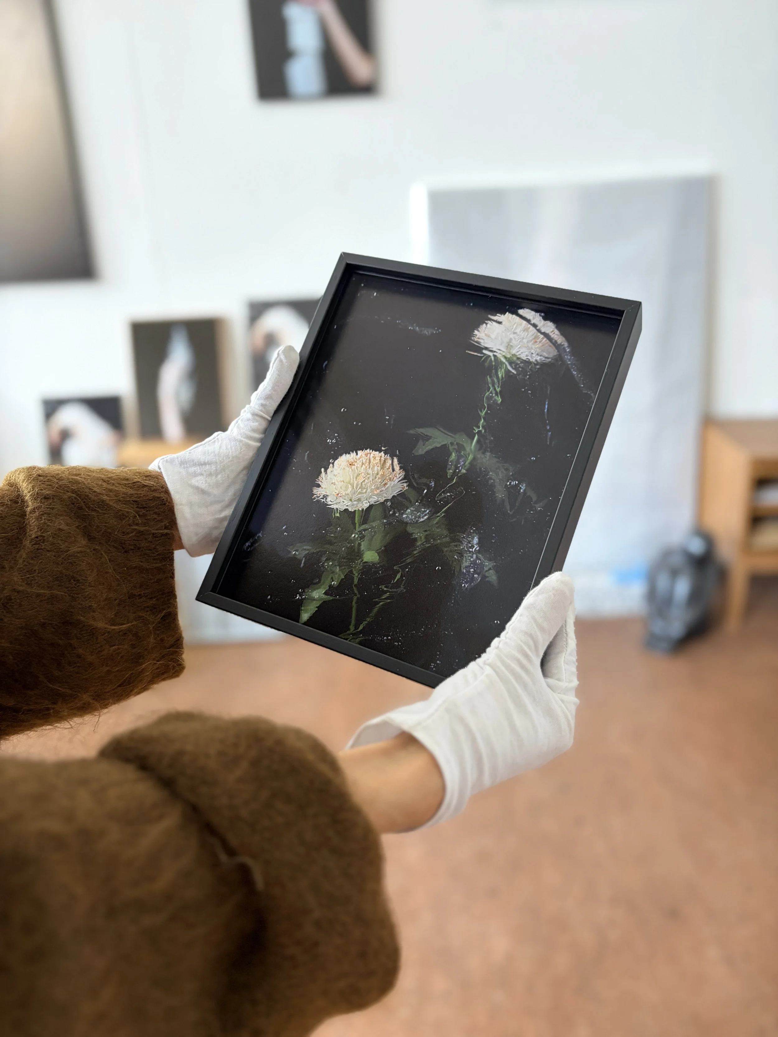 White-gloved hands hold Echoes Around the Edges by Sophie de Vos in a black frame with museum glass, with other artworks visible in her studio behind.
