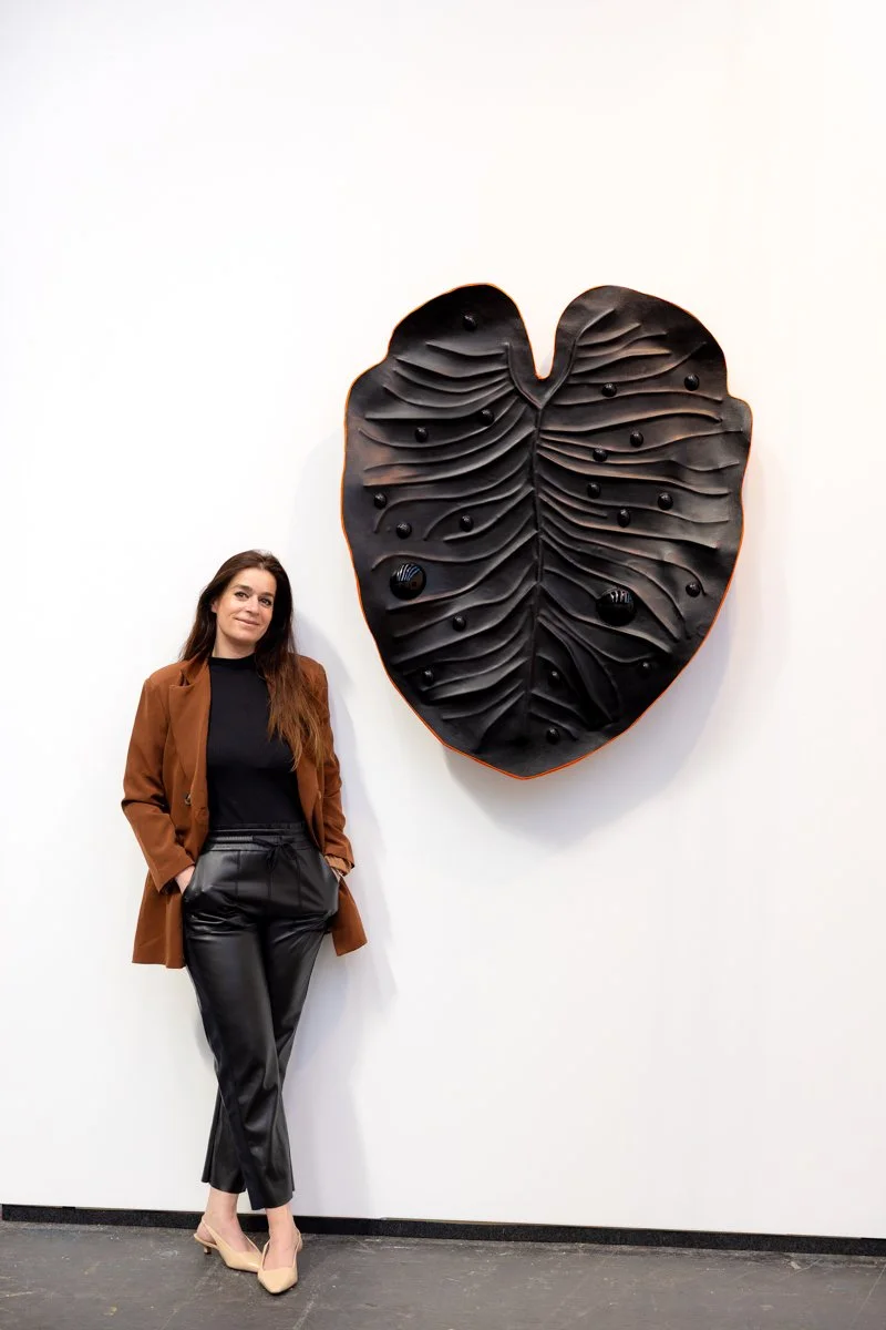 Madelon Uljee next to her large, black, leather wall sculpture in the form of a leave.