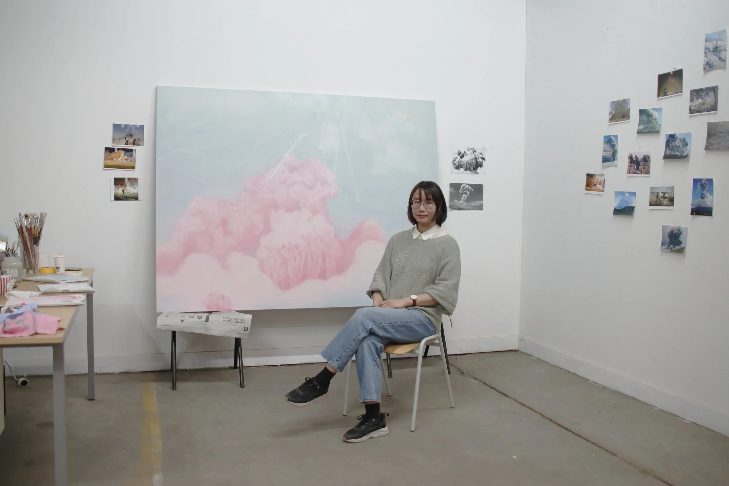Artist Mel Chan standing among her contemporary paintings in the studio, including atmospheric cloud studies exploring beauty, uncertainty and perception.
