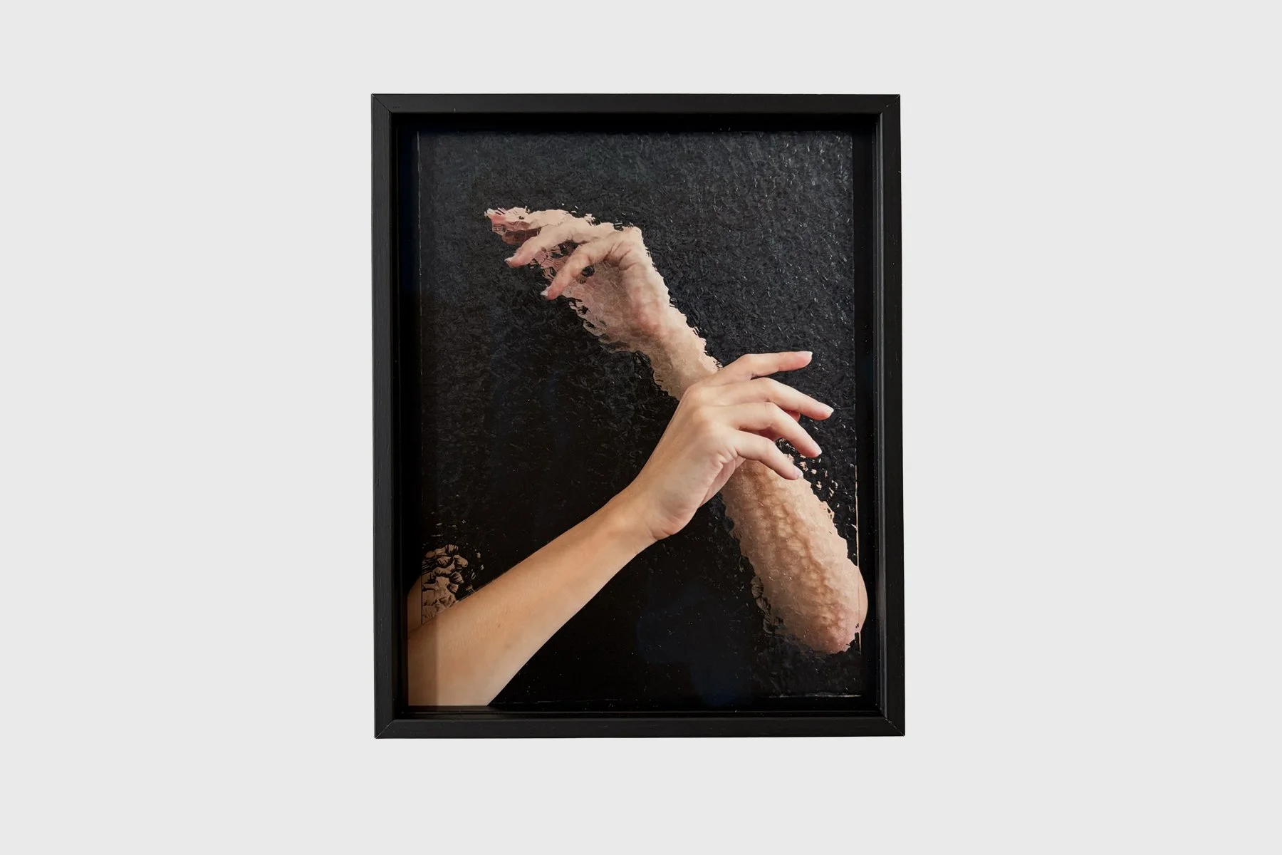 Sophie de Vos, What Lies Between: two hands and forearms cross against a dark background, one sharply defined and the other fragmented by rippled distortion. Front view in a black fram