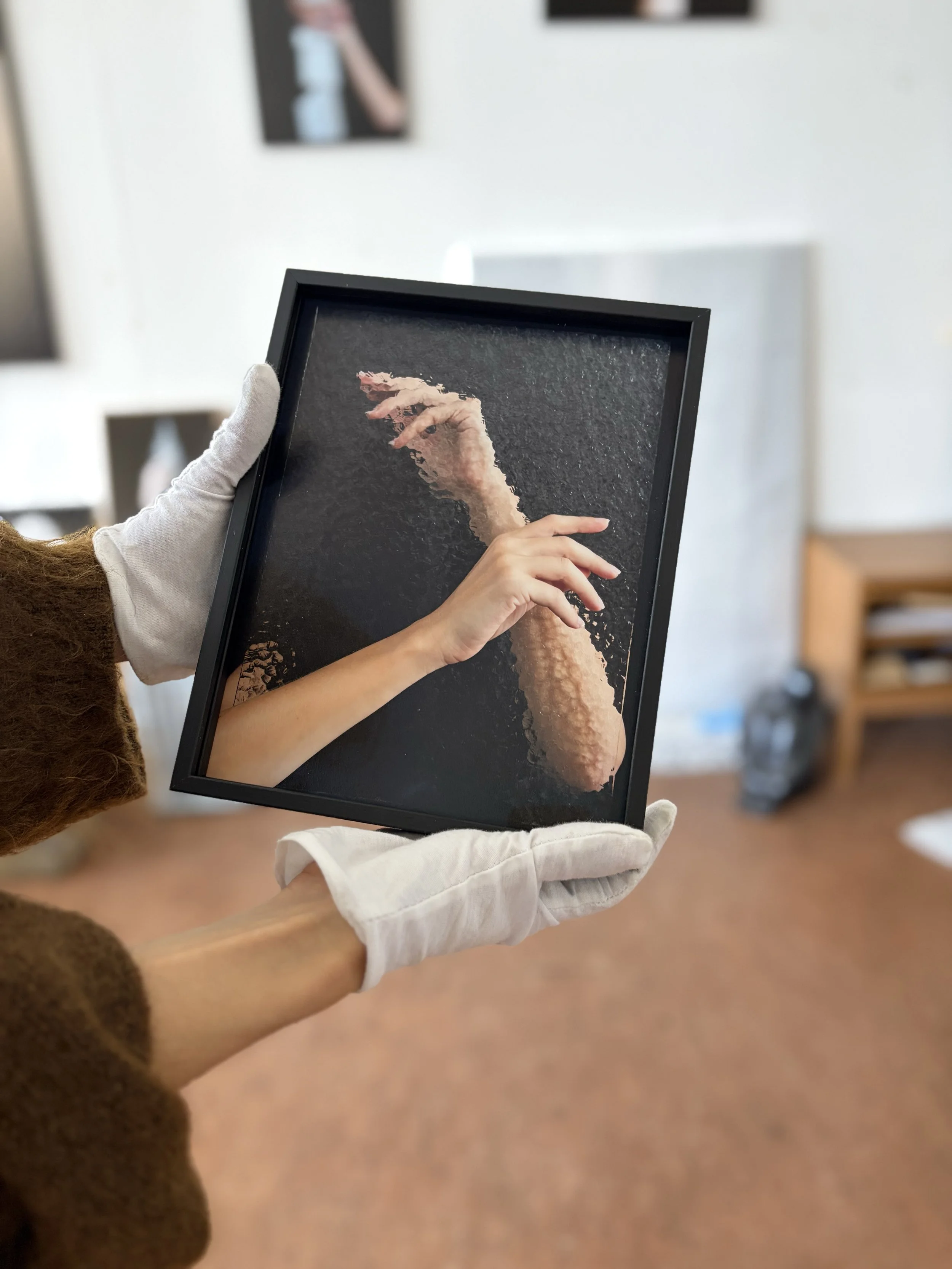 Sophie de Vos holding What Lies Between in her studio, a black-framed photographic artwork showing two hands and forearms against a dark background, with rippled distortion.