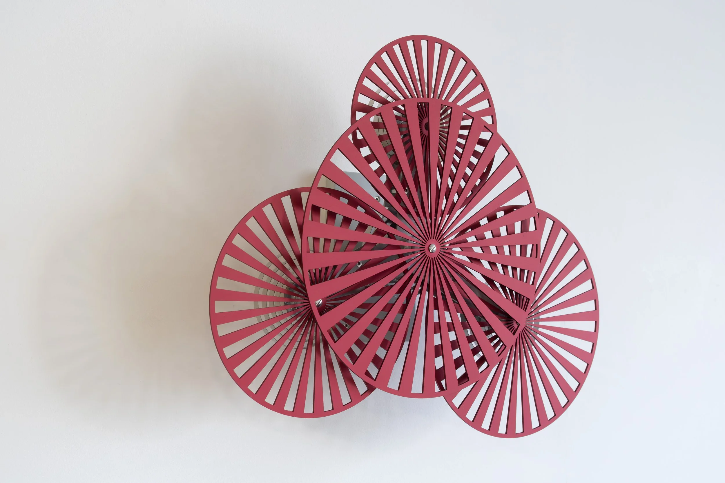 Interactive kinetic artwork by Freerk Wilbers in red anodised aluminium, featuring mechanically linked rotating circles against a white wall.