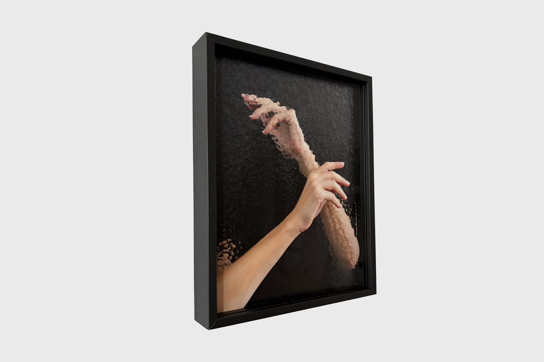 Angled view of What Lies Between by Sophie de Vos, showing its black frame and the contrast between a clearly defined hand and a distorted hand against a dark background.