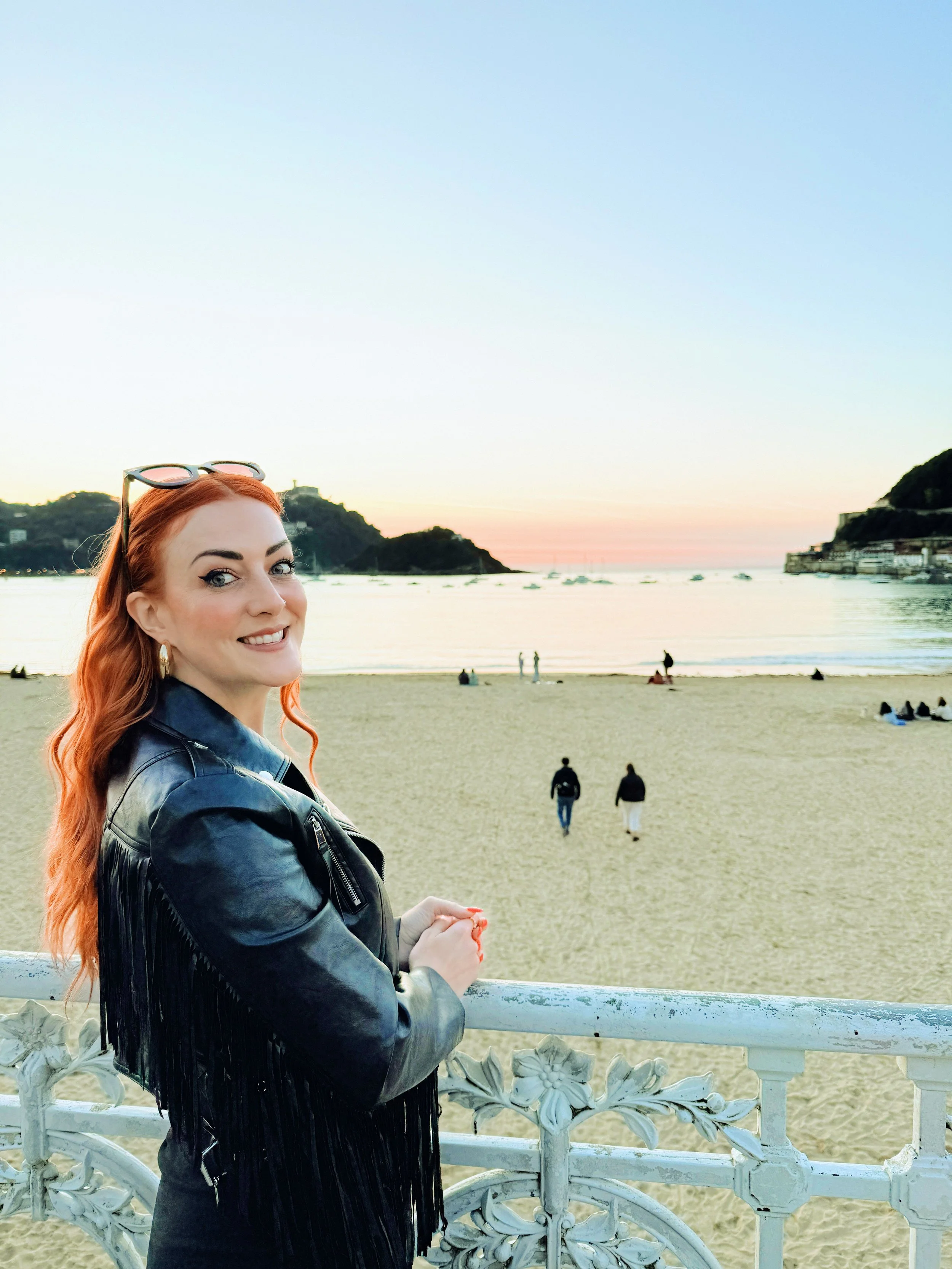 Travel and lifestyle blogger with long red hair in a black leather fringed jacket, looking out at the sunset over La Concha Beach and the bay in San Sebastian before heading into the old town for pintxos