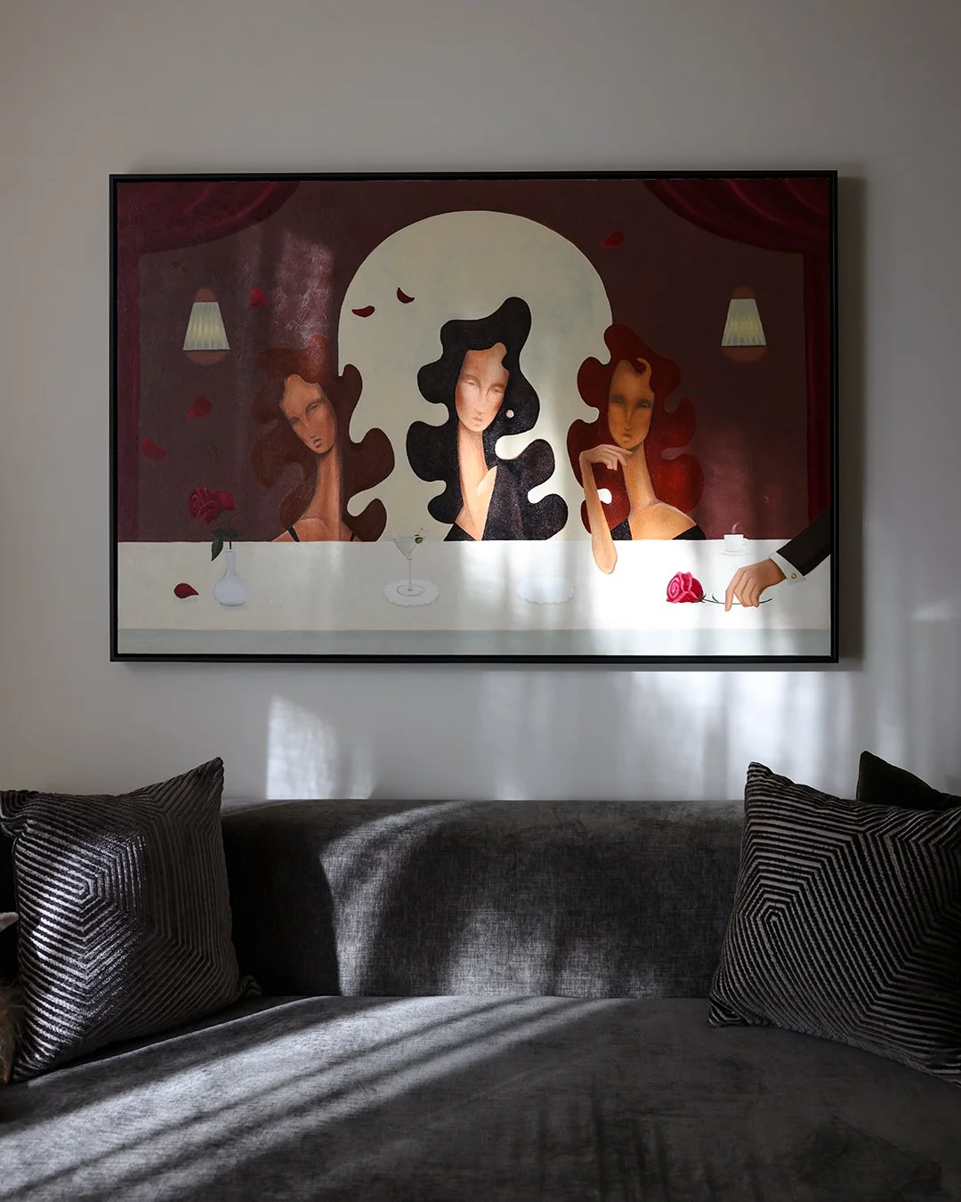 A framed artwork of three women with exaggerated flowing hair, a large moon in the background, and a hand offering a red rose on a table, hanging on a light-colored wall above a gray sofa with patterned pillows.