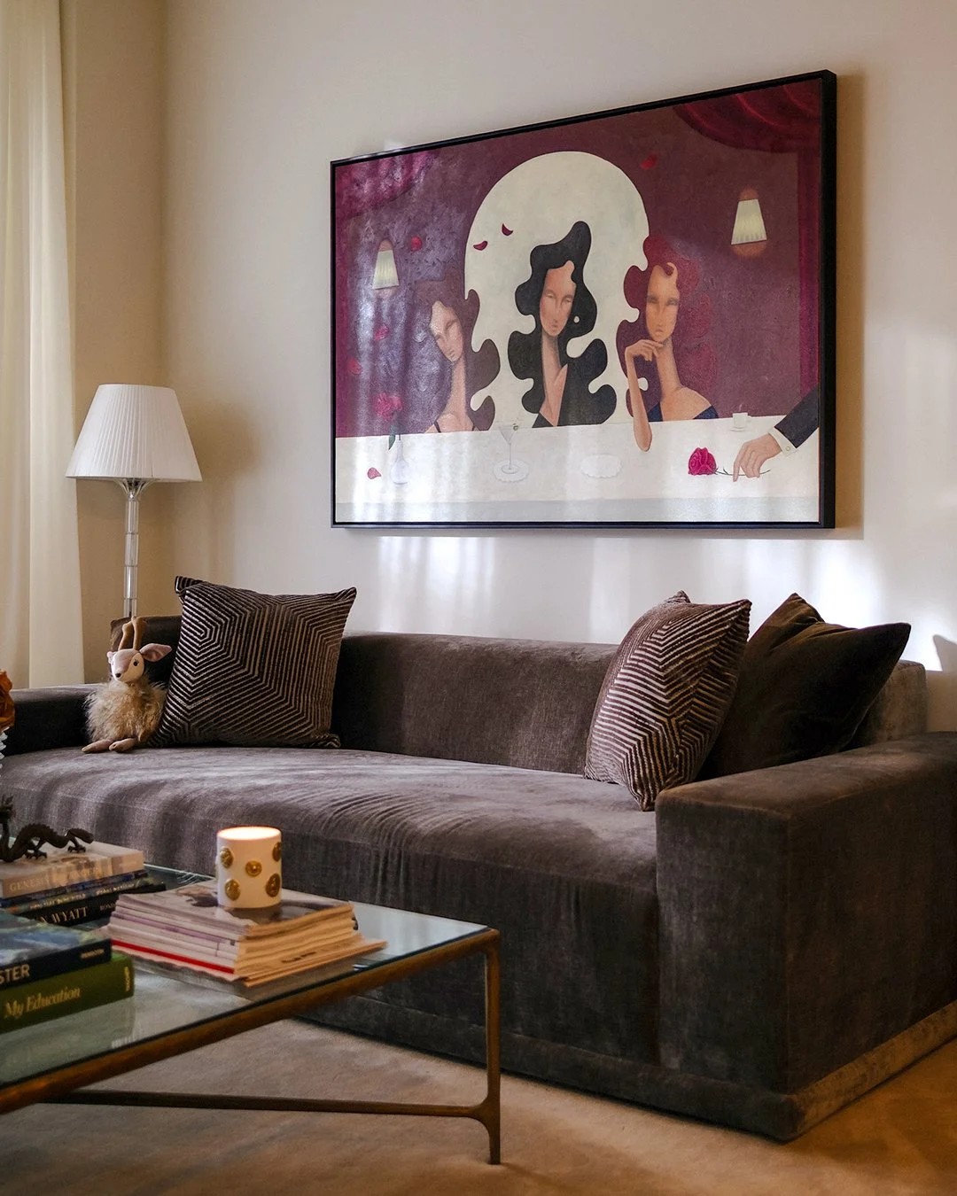 Living room with a brown velvet sofa, pillows, a side table with a candle and books, a white floor lamp, a plush toy, and a large framed artwork on the wall depicting three women at a table, with one woman holding a rose.