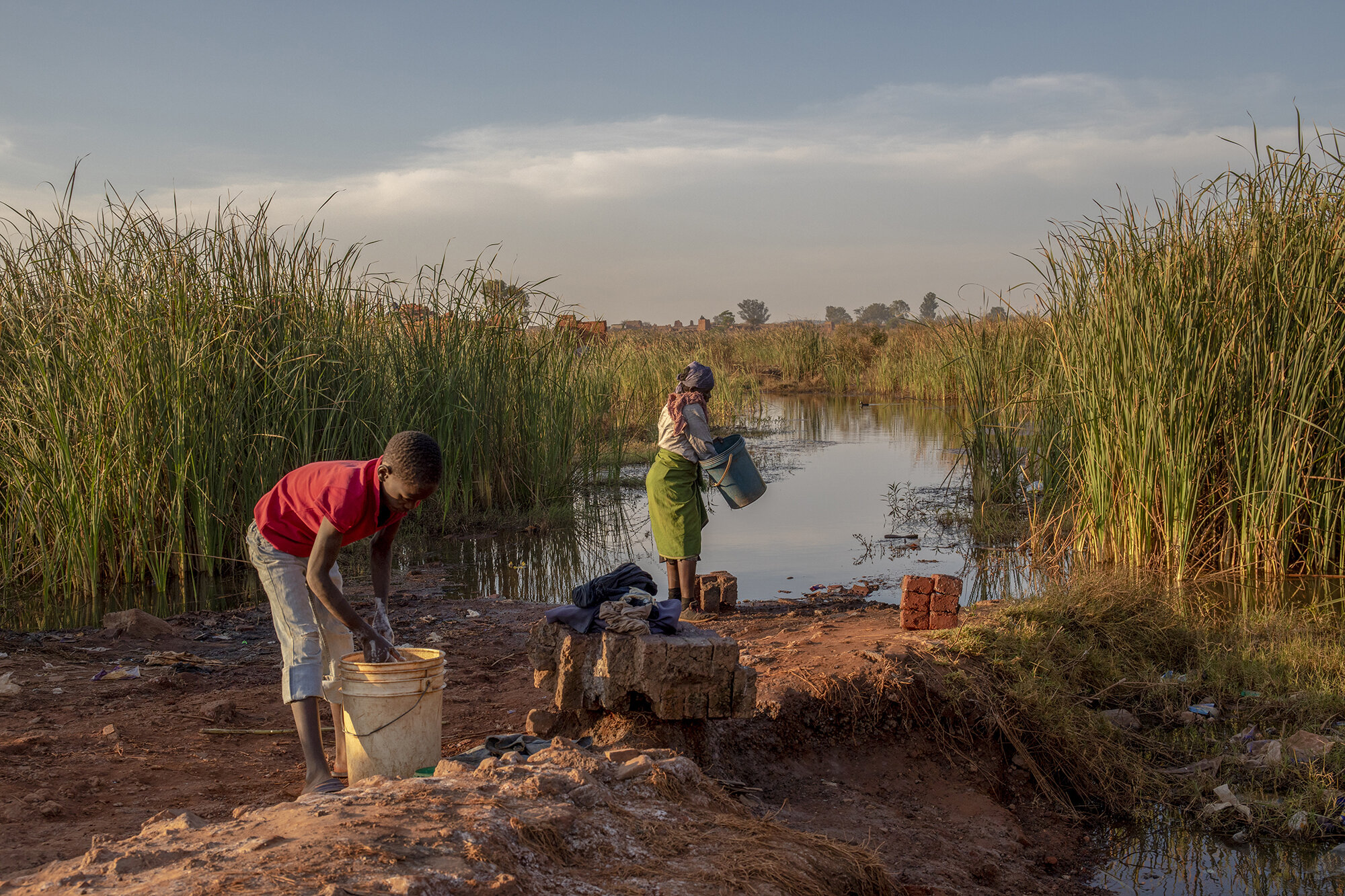  October 8, 2018: Rekina walks to a man-made dam near her home to fetch water for washing her dishes and clothes. The area is rife with informal brick makers, that have created mini-dams in the process of digging sand to make bricks. Because the wate