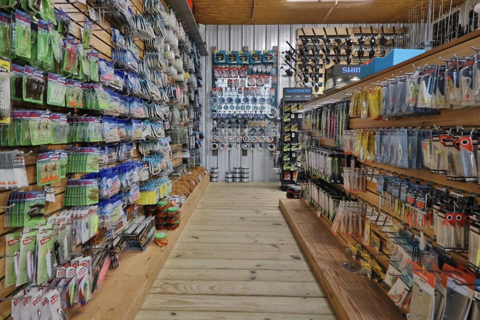 A fishing tackle shop with wooden flooring and shelves stocked with fishing gear such as lures, hooks, and reels, along with spools of fishing line and other accessories.