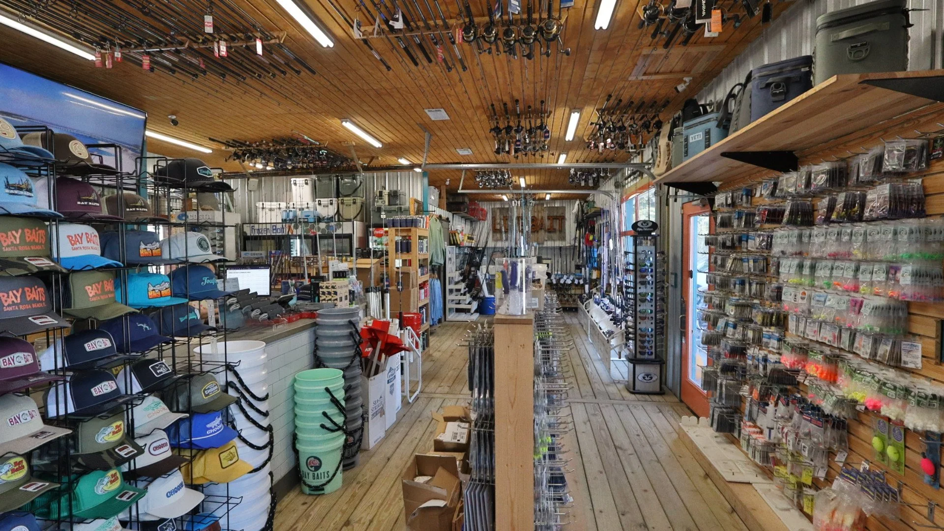 Interior of a fishing tackle shop with fishing rods hanging from the ceiling, hats on display on metal racks, and fishing lures on pegboards, with various fishing equipment and supplies arranged on shelves and counters.