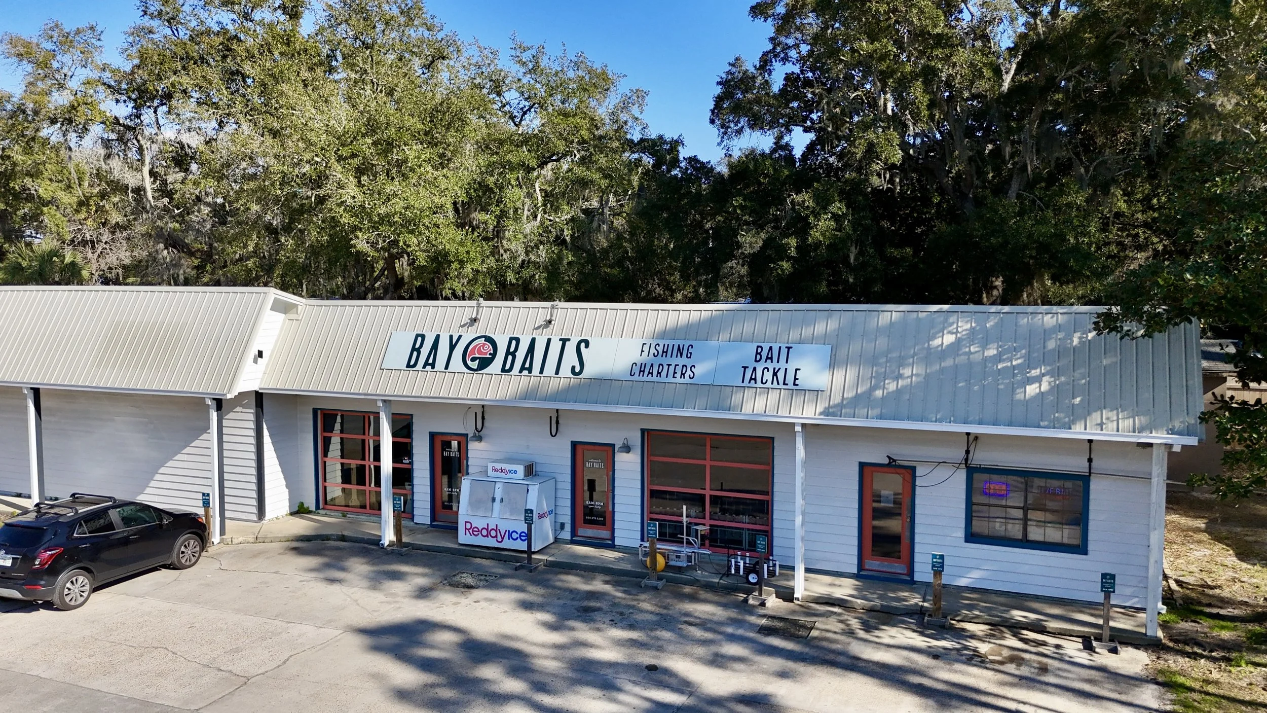 Small building with a sign that says 'BAYO BAITS' and offers fishing charters and bait tackle services. There is a Reddy Ice vending machine outside, and a parking lot with a black car parked nearby. Trees are in the background.