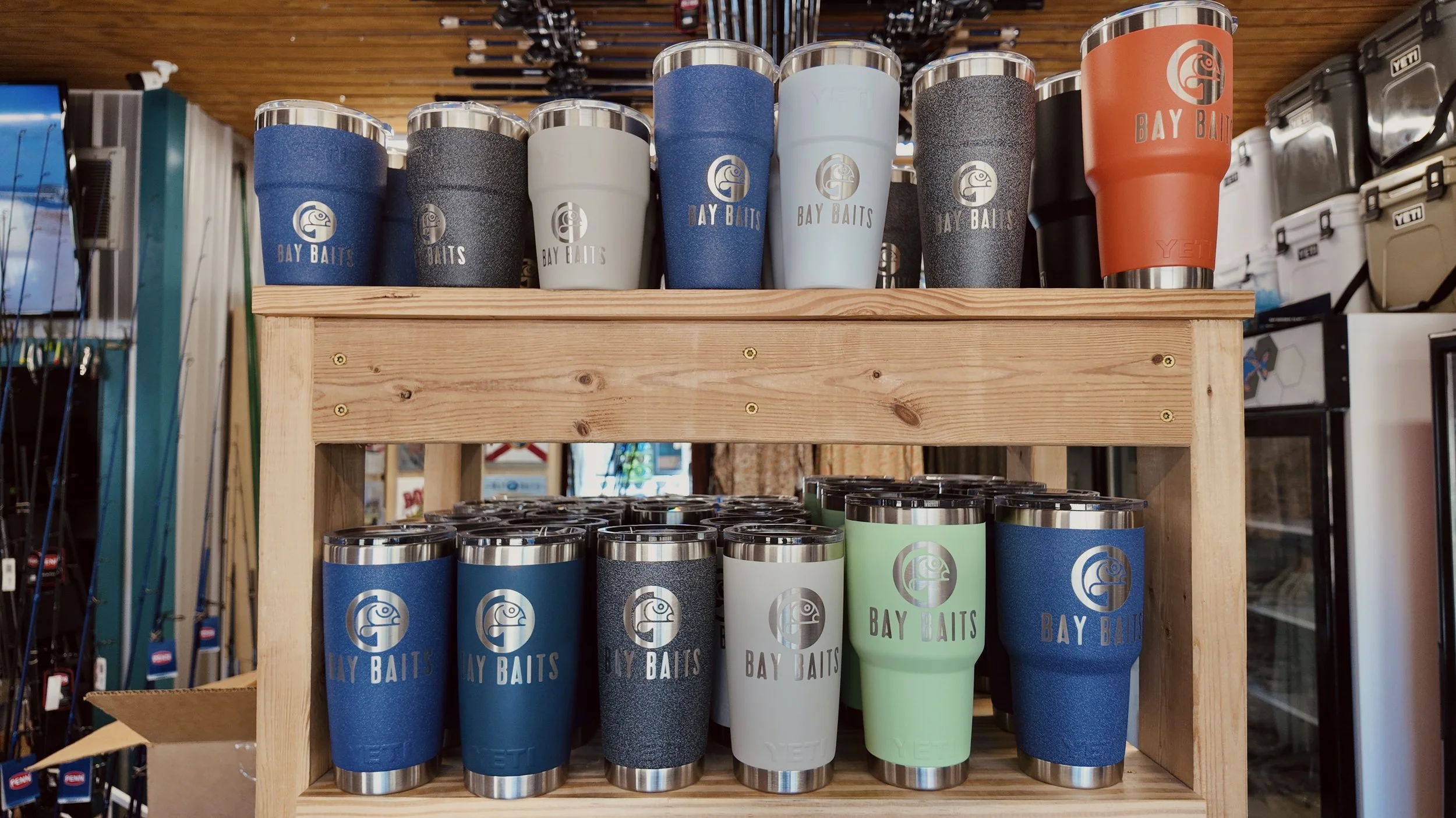 Display of insulated tumblers with logos and colors such as blue, black, white, green, and orange, on a wooden shelf at a store.