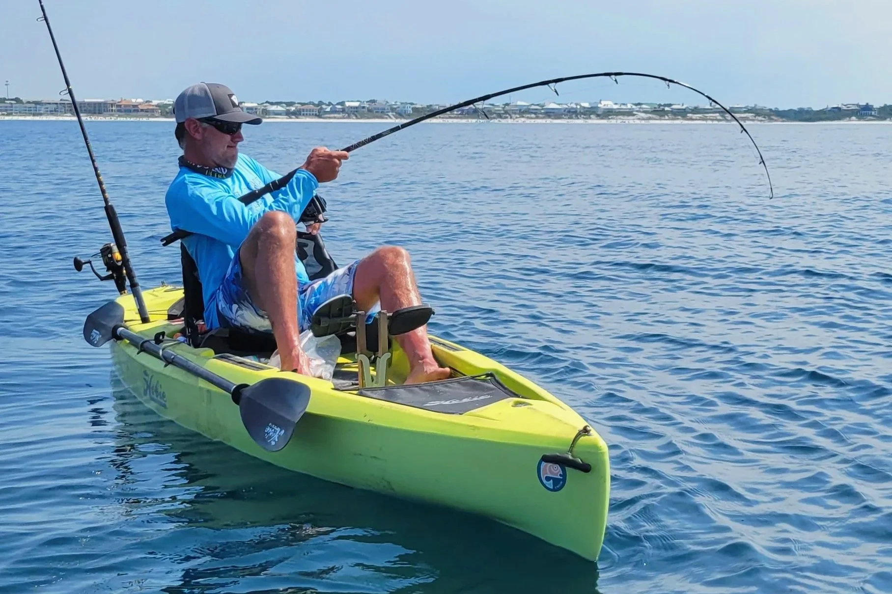 A man sitting in a yellow kayak, fishing with a rod, on calm blue water. The man is wearing a cap, sunglasses, a blue long-sleeve shirt, and shorts. In the background, there are houses and a clear sky.