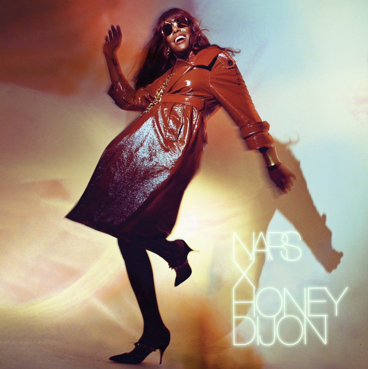 Woman with red hair wearing sunglasses, a shiny red trench coat, and black high heels, dancing against a colorful background with the text 'NARS X HONEY DIJON' in neon lights.