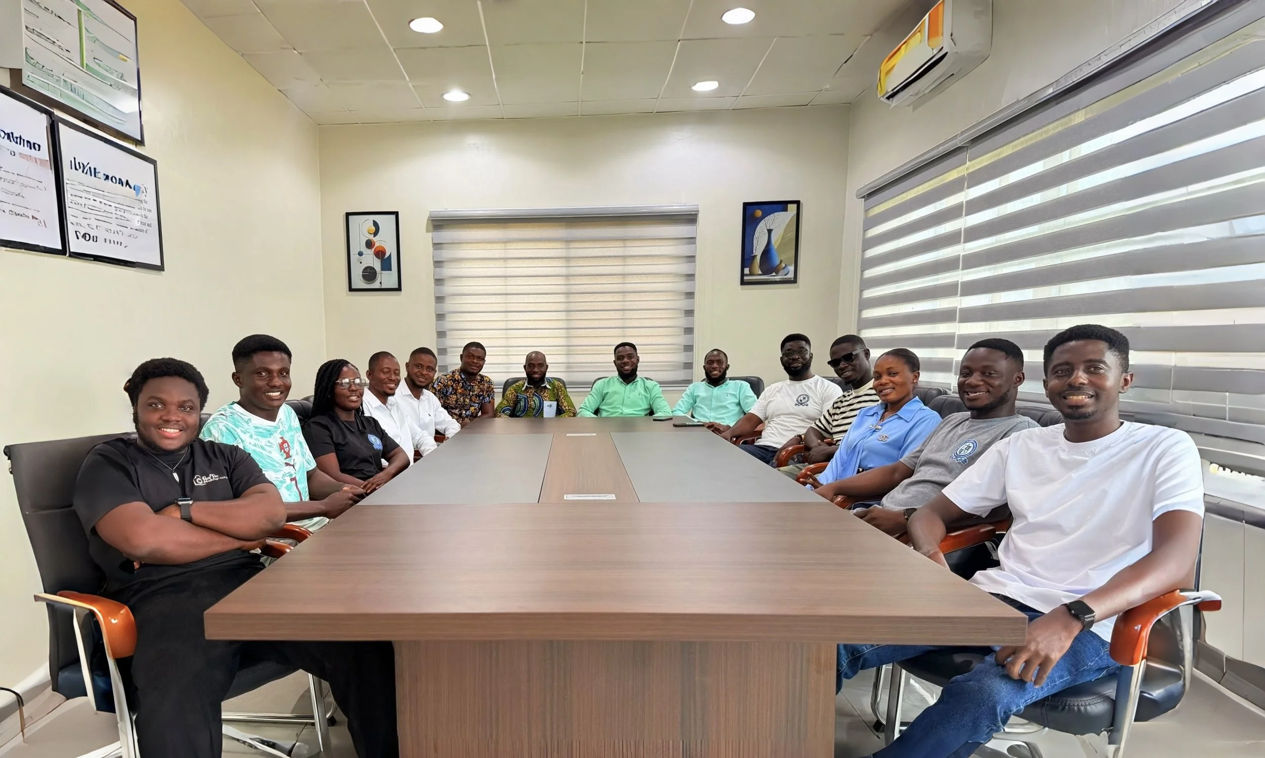 A diverse group of fifteen people sitting around a conference table in a brightly lit office meeting room, smiling at the camera.