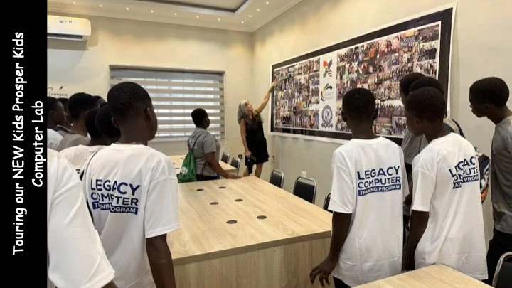 Kids in white t-shirts watching a woman present in front of a large photo collage at a computer lab.