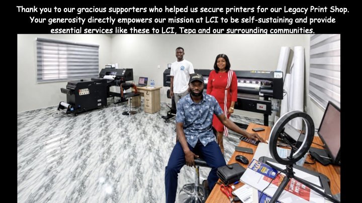 Three people inside a print shop, with large printers and printing equipment around them. One person is sitting at a desk with a computer and ring light, while two others stand behind, all smiling. The room has gray-patterned flooring and ample lighting.