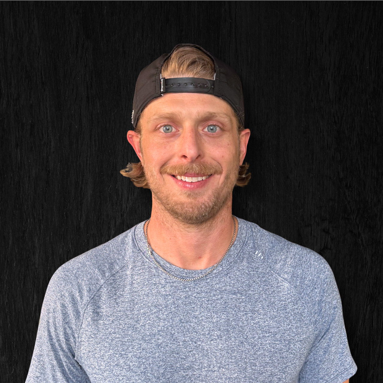 A smiling man wearing a black cap backwards and a gray athletic shirt, standing against a black textured background.
