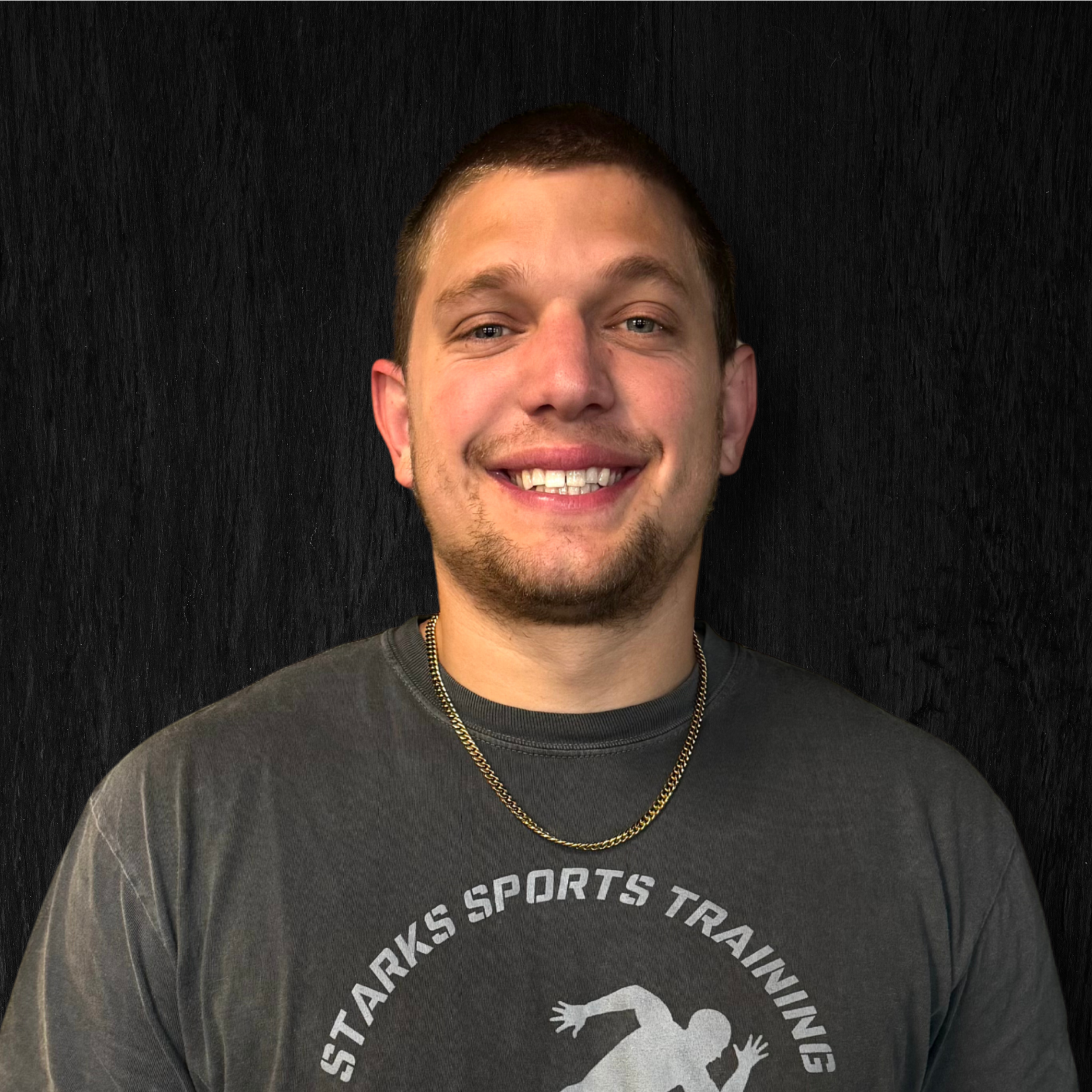 Young man smiling, wearing a gray t-shirt with a logo and gold chain necklace, standing against a black background.