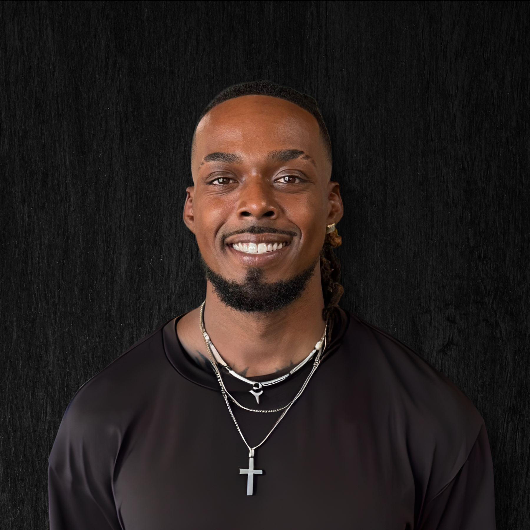 Portrait of a smiling young man with dark skin, wearing a black shirt and layered necklaces, including a cross pendant, against a black textured background.