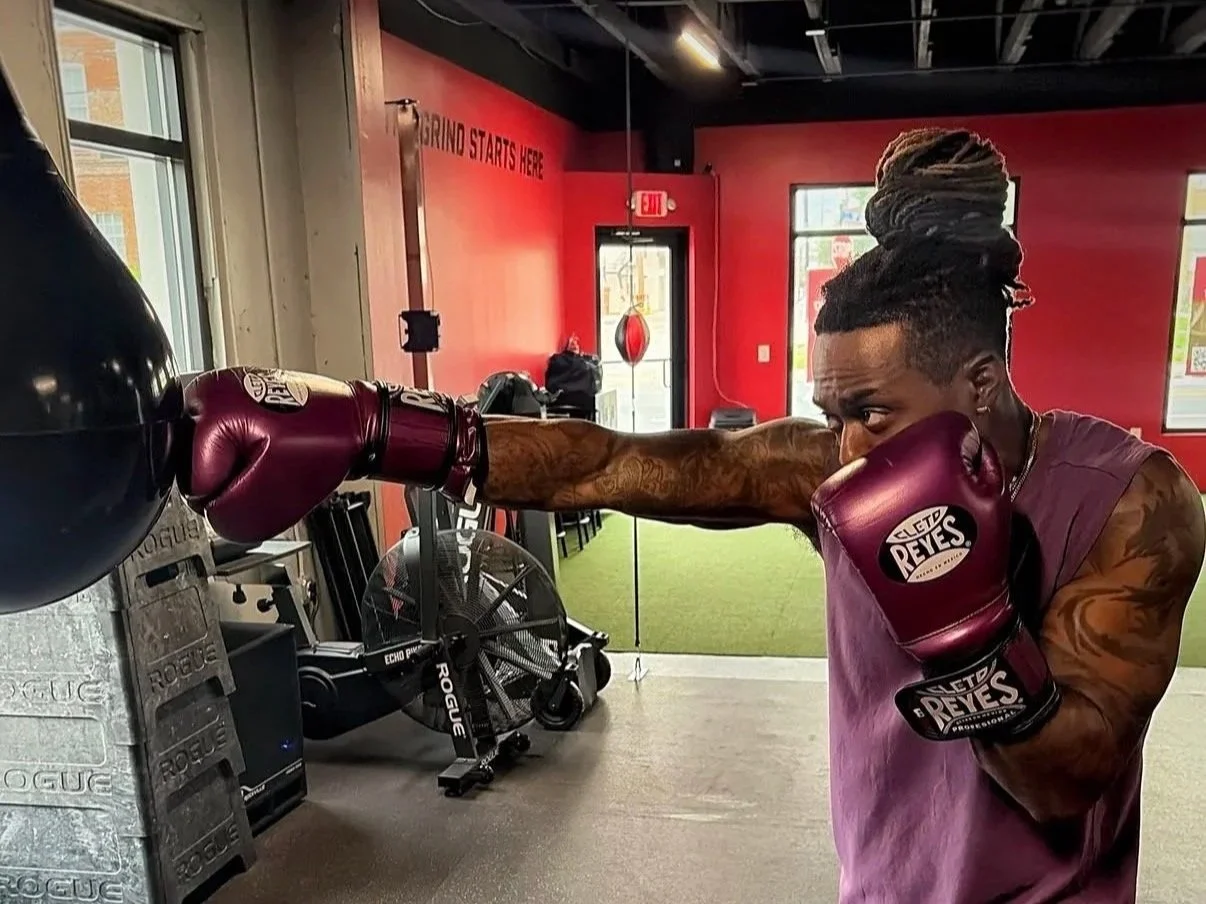 A man with styled hair and tattoos on his arm wearing boxing gloves, practicing a punch on a black punching bag in a gym with red walls and green turf flooring.