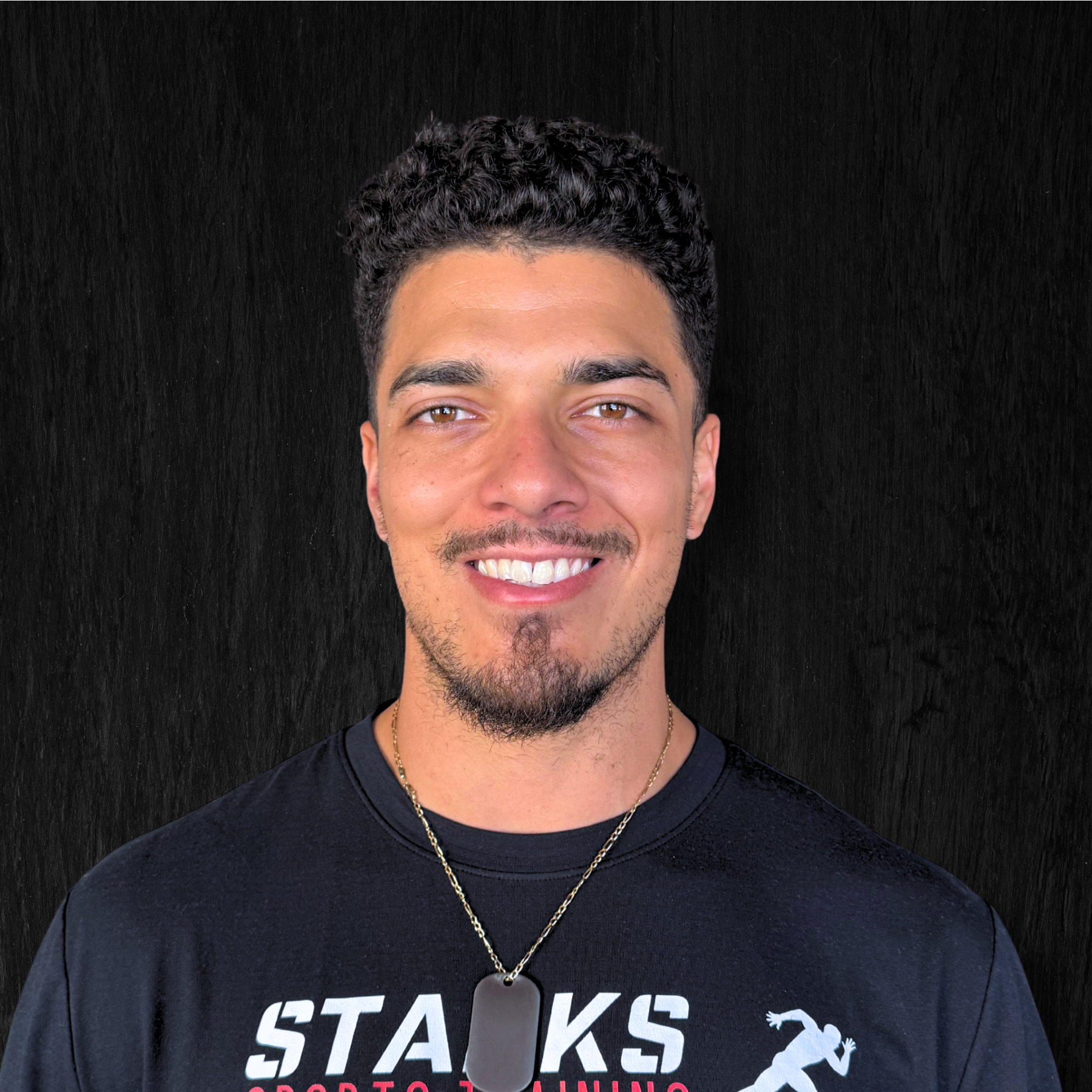 A young man with curly dark hair, light brown skin, and a goatee, smiling at the camera, wearing a black T-shirt with white lettering and a graphic of a running person, standing against a dark background.