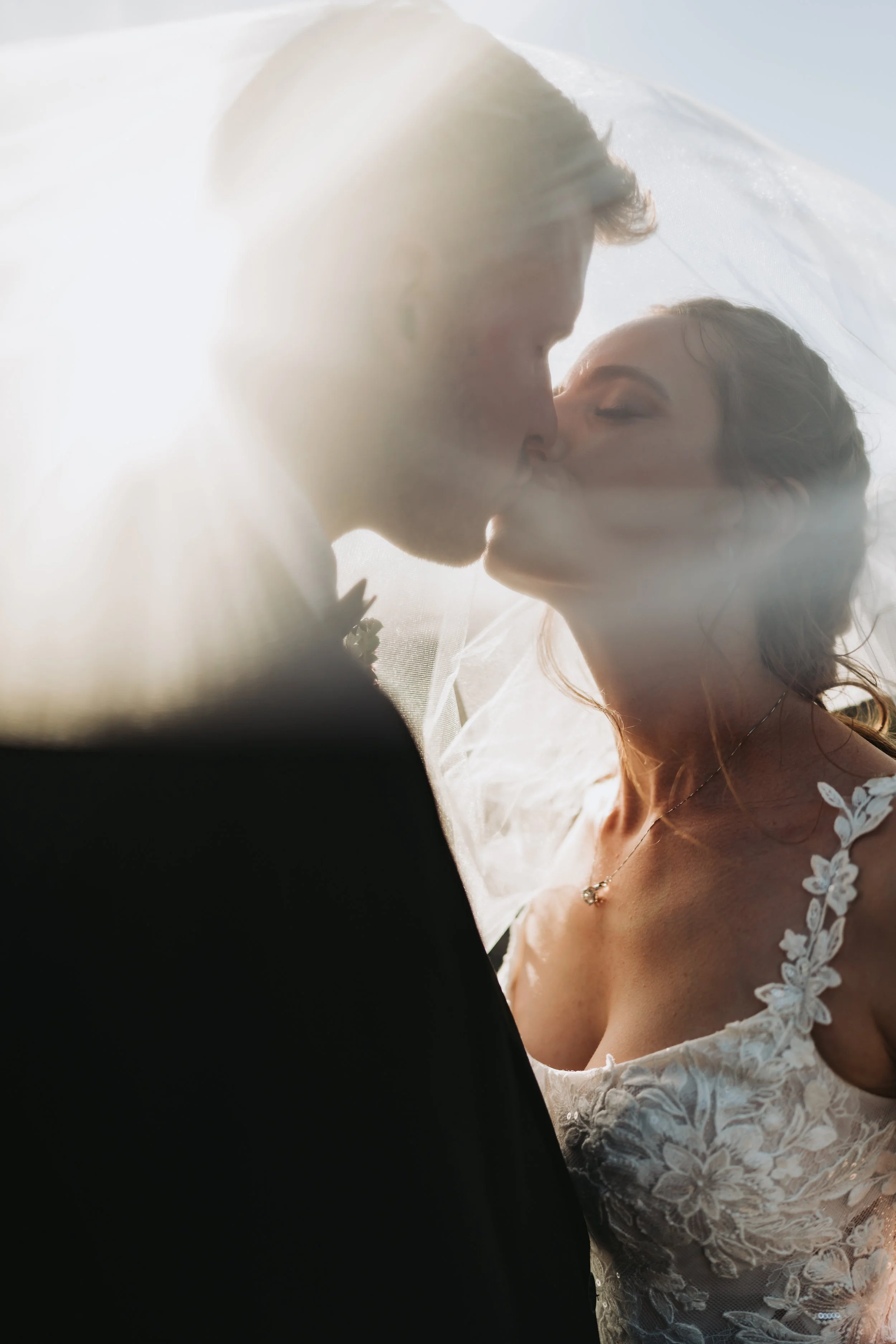 A bride and groom share a kiss with backlit sunlight creating a soft glow around them.